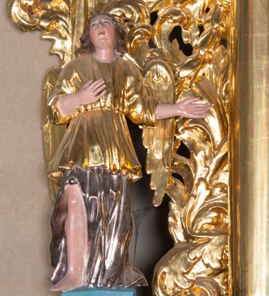 Zdjęcie nr 1: Całopostaciowa, pełnoplastyczna figura stojącego w kontrapoście anioła zwróconego na wprost. Owalną twarz o pełnych policzkach i podbródku charakteryzuje zadarty nos i zwrócone ku górze oczy. Półdługie, delikatnie kręcone włosy opadają na ramiona. Anioł prawą rękę składa na piersiach, lewą, zgiętą w łokciu kieruje w lewo. Ubrany jest w długą, srebrzoną szatę spodnią odsłaniającą prawą nogę, silnie drapowaną, o podwianym spodzie oraz krótszą, złoconą szatę wierzchnią o szerokich, sięgających łokci rękawach, przepasaną w talii. Tkanina obficie drapowana w drobne fałdki. Skrzydła złożone.