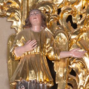 Zdjęcie nr 1: Całopostaciowa, pełnoplastyczna figura stojącego w kontrapoście anioła zwróconego na wprost. Owalną twarz o pełnych policzkach i podbródku charakteryzuje zadarty nos i zwrócone ku górze oczy. Półdługie, delikatnie kręcone włosy opadają na ramiona. Anioł prawą rękę składa na piersiach, lewą, zgiętą w łokciu kieruje w lewo. Ubrany jest w długą, srebrzoną szatę spodnią odsłaniającą prawą nogę, silnie drapowaną, o podwianym spodzie oraz krótszą, złoconą szatę wierzchnią o szerokich, sięgających łokci rękawach, przepasaną w talii. Tkanina obficie drapowana w drobne fałdki. Skrzydła złożone.