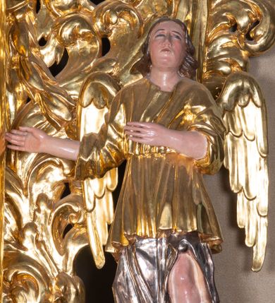 Zdjęcie nr 1: Całopostaciowa, pełnoplastyczna figura stojącego w kontrapoście anioła zwróconego na wprost. Owalną twarz o pełnych policzkach i podbródku charakteryzuje zadarty nos i zwrócone ku górze oczy. Półdługie, delikatnie kręcone włosy opadają na ramiona. Anioł obydwie, zgięte w łokciach ręce kieruje w prawo. Ubrany jest w długą, srebrzoną szatę spodnią odsłaniającą lewą nogę, silnie drapowaną, o podwianym spodzie oraz krótszą, złoconą szatę wierzchnią o szerokich, sięgających łokci rękawach, przepasaną w talii. Tkanina obficie drapowana w drobne fałdki. Skrzydła złożone.