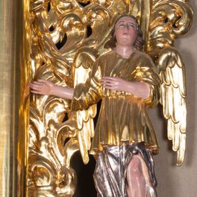 Zdjęcie nr 1: Całopostaciowa, pełnoplastyczna figura stojącego w kontrapoście anioła zwróconego na wprost. Owalną twarz o pełnych policzkach i podbródku charakteryzuje zadarty nos i zwrócone ku górze oczy. Półdługie, delikatnie kręcone włosy opadają na ramiona. Anioł obydwie, zgięte w łokciach ręce kieruje w prawo. Ubrany jest w długą, srebrzoną szatę spodnią odsłaniającą lewą nogę, silnie drapowaną, o podwianym spodzie oraz krótszą, złoconą szatę wierzchnią o szerokich, sięgających łokci rękawach, przepasaną w talii. Tkanina obficie drapowana w drobne fałdki. Skrzydła złożone.