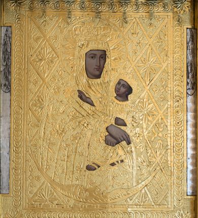 Zdjęcie nr 1: Obraz w kształcie stojącego prostokąta z przedstawieniem Matki Boskiej z Dzieciątkiem ukazanej w półpostaci, nieznacznie zwróconej w lewo. Owalną twarz charakteryzują wyraziste, delikatnie modelowane rysy o dużych oczach, długim nosie i wydatnych ustach. Rękami o skrzyżowanych dłoniach, z wyprostowanymi palcami prawej ręki, podtrzymuje siedzące na jej lewym ramieniu Dzieciątko. Jezus zwrócony jest w trzech czwartych w prawo. Lekko odchyloną głowę charakteryzuje owalna twarz o  wyrazistych, delikatnie modelowanych rysach o dużych oczach, długim nosie i wydatnych ustach. Dzieciątko prawą rękę wyciąga przed siebie w geście błogosławieństwa, lewą podtrzymuje zamkniętą księgę. Obraz pokryty jest metalową blachą, w której zaznaczony jest zarys ubioru postaci. Maria ma na sobie suknię i osłaniający szczelnie sylwetkę, nałożony na głowę płaszcz. Jezus ubrany jest w długą suknię. Szaty zdobi ornamentyka roślinna z motywami gwiazd. Głowy postaci w koronach zamkniętych okalają nimby z promienistą glorią w środku. Tło obrazu pokrywa motyw romboidalnej kraty z motywami rozet.  U dołu postaci wąski sierp księżyca. Przedstawienie ujęte pasami ornamentu cekinowego z rozetami w narożach.      
