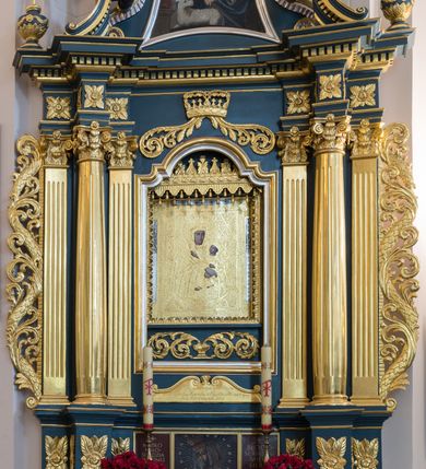 Zdjęcie nr 2: Obraz w kształcie stojącego prostokąta z przedstawieniem Matki Boskiej z Dzieciątkiem ukazanej w półpostaci, nieznacznie zwróconej w lewo. Owalną twarz charakteryzują wyraziste, delikatnie modelowane rysy o dużych oczach, długim nosie i wydatnych ustach. Rękami o skrzyżowanych dłoniach, z wyprostowanymi palcami prawej ręki, podtrzymuje siedzące na jej lewym ramieniu Dzieciątko. Jezus zwrócony jest w trzech czwartych w prawo. Lekko odchyloną głowę charakteryzuje owalna twarz o  wyrazistych, delikatnie modelowanych rysach o dużych oczach, długim nosie i wydatnych ustach. Dzieciątko prawą rękę wyciąga przed siebie w geście błogosławieństwa, lewą podtrzymuje zamkniętą księgę. Obraz pokryty jest metalową blachą, w której zaznaczony jest zarys ubioru postaci. Maria ma na sobie suknię i osłaniający szczelnie sylwetkę, nałożony na głowę płaszcz. Jezus ubrany jest w długą suknię. Szaty zdobi ornamentyka roślinna z motywami gwiazd. Głowy postaci w koronach zamkniętych okalają nimby z promienistą glorią w środku. Tło obrazu pokrywa motyw romboidalnej kraty z motywami rozet.  U dołu postaci wąski sierp księżyca. Przedstawienie ujęte pasami ornamentu cekinowego z rozetami w narożach.      
