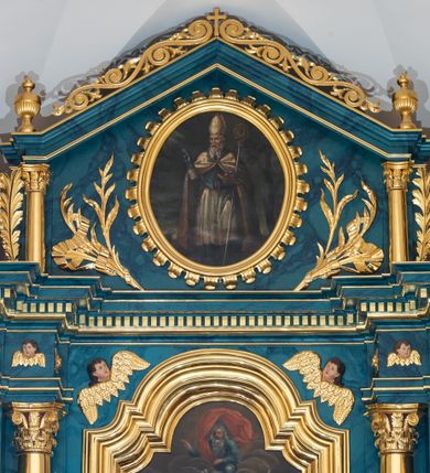 Zdjęcie nr 11: Ołtarz przyścienny, jednokondygnacyjny, jednoosiowy, ze zwieńczeniem. Ołtarz prostopadłościenny, tabernakulum z tronem wystawienia w formie architektonicznej na rzucie trapezoidalnym. W przedniej ściance wysokiego cokołu, o narożach ujętych spływami wolutowymi, są drzwiczki w kształcie prostokąta zamkniętego półkolistym łukiem nadwieszonym zdobione płaskorzeźbionym przedstawieniem kielicha eucharystycznego. Cokół wieńczy gzyms przekształcający się pośrodku w parę stykających się wolut. Na występach nad spływami cokołu osadzone kolumienki wspierające wyłamujące się ponad nimi belkowanie baldachimu tronu wystawienia. Zwieńczenie w formie pary stykających się spływów wolutowych z motywem paludamentu. Zaplecek zdobiony płycinami z ornamentalnymi obramieniami z motywami rocaille’u, w górnej partii płaskorzeźbiony trójkąt z okiem Opatrzności Bożej ujęty promienistą glorią. W zwieńczeniu para wazonów i para figurek aniołków, na szczycie księga z siedmioma pieczęciami i leżącym barankiem paschalnym.  
Retabulum osadzone na zdwojonym cokole, w górnej partii o zryzalitowanych bokach. Część środkową ujmują trzy pary kolumn, z których środkowe wysunięte, wspierających przerwane w partii architrawu i fryzu belkowanie. Zwieńczenie ujęte parą kolumn wspierających gzyms w formie trójkątnego przyczółka. Pola cokołu retabulum wypełnia stylizowana ornamentyka roślinna, partie fryzu oraz górne naroża części środkowej zdobią uskrzydlone główki anielskie, a gzyms kostkowanie. Zwieńczenie ujęte po bokach motywem liści paproci, pole ulistnionymi gałązkami z wstążką. Krawędź gzymsu wieńczącego zdobi wić roślinna z krzyżem pośrodku, po bokach para wazonów. Całość ujmuje para figur klęczących aniołów. Pole części środkowej w kształcie stojącego prostokąta ujętego profilowaną, złoconą ramą z przedstawieniem Narodzenia Najświętszej Marii Panny. W zwieńczeniu obraz w kształcie stojącego owalu, ujętego ramą zdobioną akantowymi liśćmi, z wizerunkiem św. Mikołaja. Struktura marmoryzowana w odcieniach błękitów, detal architektoniczny i ornamentyka złocone.   
