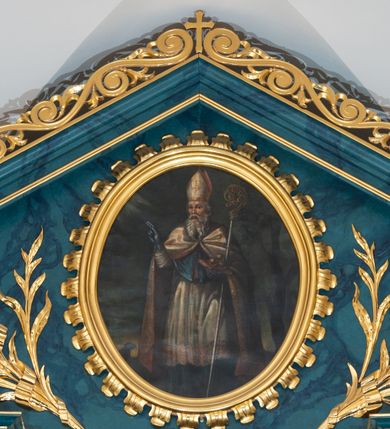 Zdjęcie nr 15: Ołtarz przyścienny, jednokondygnacyjny, jednoosiowy, ze zwieńczeniem. Ołtarz prostopadłościenny, tabernakulum z tronem wystawienia w formie architektonicznej na rzucie trapezoidalnym. W przedniej ściance wysokiego cokołu, o narożach ujętych spływami wolutowymi, są drzwiczki w kształcie prostokąta zamkniętego półkolistym łukiem nadwieszonym zdobione płaskorzeźbionym przedstawieniem kielicha eucharystycznego. Cokół wieńczy gzyms przekształcający się pośrodku w parę stykających się wolut. Na występach nad spływami cokołu osadzone kolumienki wspierające wyłamujące się ponad nimi belkowanie baldachimu tronu wystawienia. Zwieńczenie w formie pary stykających się spływów wolutowych z motywem paludamentu. Zaplecek zdobiony płycinami z ornamentalnymi obramieniami z motywami rocaille’u, w górnej partii płaskorzeźbiony trójkąt z okiem Opatrzności Bożej ujęty promienistą glorią. W zwieńczeniu para wazonów i para figurek aniołków, na szczycie księga z siedmioma pieczęciami i leżącym barankiem paschalnym.  
Retabulum osadzone na zdwojonym cokole, w górnej partii o zryzalitowanych bokach. Część środkową ujmują trzy pary kolumn, z których środkowe wysunięte, wspierających przerwane w partii architrawu i fryzu belkowanie. Zwieńczenie ujęte parą kolumn wspierających gzyms w formie trójkątnego przyczółka. Pola cokołu retabulum wypełnia stylizowana ornamentyka roślinna, partie fryzu oraz górne naroża części środkowej zdobią uskrzydlone główki anielskie, a gzyms kostkowanie. Zwieńczenie ujęte po bokach motywem liści paproci, pole ulistnionymi gałązkami z wstążką. Krawędź gzymsu wieńczącego zdobi wić roślinna z krzyżem pośrodku, po bokach para wazonów. Całość ujmuje para figur klęczących aniołów. Pole części środkowej w kształcie stojącego prostokąta ujętego profilowaną, złoconą ramą z przedstawieniem Narodzenia Najświętszej Marii Panny. W zwieńczeniu obraz w kształcie stojącego owalu, ujętego ramą zdobioną akantowymi liśćmi, z wizerunkiem św. Mikołaja. Struktura marmoryzowana w odcieniach błękitów, detal architektoniczny i ornamentyka złocone.   
