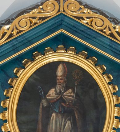 Zdjęcie nr 1: Obraz w kształcie stojącego owalu ujęty profilowaną, złoconą ramą o krawędzi zdobionej liśćmi akantu. Malowidło przedstawia reprezentacyjny, całopostaciowy wizerunek św. Mikołaja stojącego w lekkim kontrapoście i zwróconego na wprost. Skierowaną w trzech czwartych w prawo głowę charakteryzują rysy mężczyzny w podeszłym wieku, z długim, siwym zarostem. Święty Mikołaj jest ubrany w strój pontyfikalny składający się z ciemnoniebieskiej sutanny, rokiety, ciemnoniebieskiego mantoletu oraz białej kapy z czerwonym podszyciem, zdobionej złotym haftem. Na głowie ma białą infułę, na dłoniach niebieskie rękawiczki. Święty prawą rękę unosi w geście błogosławieństwa, w lewej, podtrzymując pastorał, ma zamkniętą księgę z leżącymi na niej trzema złotymi kulami. Postać ukazana jest na tle pejzażu z obniżoną linią horyzontu, po prawej stronie widoczne jest duże drzewo. Niebo pokrywają ciemne chmury, w lewej górnej części kompozycji widoczna jest wiązka promieni.     