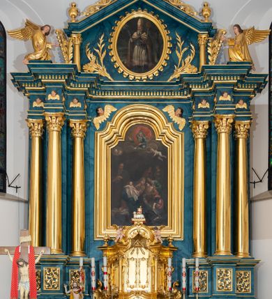 Zdjęcie nr 2: Obraz jest w kształcie stojącego prostokąta zamkniętego nadwieszonym łukiem z uskokiem, ujętego profilowaną, złoconą ramą. Dwustrefowa kompozycja przedstawia scenę Narodzin Marii. W dolnej partii, pośrodku ukazana jest siedząca i zwrócona w trzech czwartych w prawo św. Anna. Odchylając się znacznie, głowę kieruje w lewo. Święta ubrana jest w jasnoczerwoną suknię, ciemnoniebieski płaszcz i ciemnożółtą chustę osłaniającą jej głowę i ramiona. Na stopach ma sandały. Oburącz podtrzymuje małą Marię ukazaną w pozycji siedzącej, zwróconą nieznacznie w lewo, z prawą rączką wyciągniętą w bok, lewą wspartą na kolanie. Jej nagą sylwetkę osłania obszerna, biała szata. Święta Anna pochyla się ku przyklękającej obok kobiecie zwróconej w trzech czwartych w prawo. Jest ona ubrana w ciemnoniebieską bluzkę i zieloną spódnicę, które są widoczne spod czerwonej sukni wierzchniej. Głęboki dekolt okala żółty szal, a głowę kobiety osłania ciemna chusta. Niewiasta w lewej ręce trzyma srebrny dzban, w prawej powijaki. Tuż za nią stoi pochylona ku św. Annie druga z kobiet, wspierając się ręką na ramieniu pierwszej. Ubrana jest w zieloną suknię, w upiętych włosach ma niebieską ozdobę. W głębi widoczne są pozostałe dwie, zwrócone ku sobie kobiety. Stojąca po prawej stronie, ubrana w niebieską suknię i chustę na głowie, prawą rękę wspiera na piersiach, lewą na brzuchu. Druga ma na sobie ciemną suknię, czerwony płaszcz i chustę na głowie. Zaokrąglone twarze kobiet i Marii charakteryzują pełne rysy. Po lewej stronie kompozycji, tuż za św. Anną stoi św. Joachim w trzech czwartych zwrócony w lewo. Pociągłą twarz mężczyzny charakteryzują wyraziste rysy i długi, siwy zarost. Ubrany jest w ciemnobrązową suknię i ciemnozielony płaszcz. Ręce, z trzymaną w prawej dłoni księgą, skład na klatce piersiowej. Na pierwszym planie, u stóp św. Anny leży miedziana misa. W górnej partii obrazu pośród obłoków ukazana jest zwrócona w trzech czwartych w prawo sylwetka spoglądającego w dół Boga Ojca o długich, siwych włosach i zaroście. Ubrany jest w niebieską suknię i czerwony, silnie rozwiany płaszcz. Prawą rękę unosi w geście błogosławieństwa, w lewej, wspartej na globie, trzyma berło. Głowę Boga Ojca ujmuje trójkątny nimb. Poniżej ukazana jest gołębica Ducha Świętego. Na tle obłoków unosi się para uskrzydlonych główek anielskich po lewej stronie kompozycji i anioł po prawej, o nagiej sylwetce przesłoniętej niebieską szatą. W wyciągniętej przed siebie ręce trzyma koronę. 
Postaci ukazane są na ciemnym tle w odcieniach szarości. Kolorystyka obrazu jest ciemna, dominują zgaszone barwy delikatnie modelowane światłocieniowo.  
