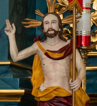 Zdjęcie nr 1: Całopostaciowa, pełnoplastyczna figura przedstawia Chrystusa Zmartwychwstałego zwróconego na wprost i stojącego w kontrapoście na czworobocznej, profilowanej podstawie. Owalną twarz charakteryzują delikatne rysy i krótki zarost. Długie, brązowe włosy opadają na ramiona. Nagie ciało Jezusa o  uwydatnionej muskulaturze i podkreślonej linii żeber osłania białe perizonium przewiązane na biodrach i spięty na prawym ramieniu obszerny, rozwiany czerwony płaszcz o żółtym podszyciu. Na dłoniach, stopach i prawym boku widoczne ślady ran. Prawa, zgięta i odchylona w bok ręka jest uniesiona w geście błogosławieństwa, lewą Jezus podtrzymuje złocony drzewiec czerwonej chorągwi. Głowę okalają wiązki złotych promieni. 
