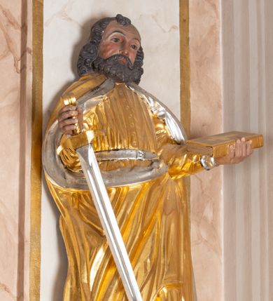 Zdjęcie nr 1: Całopostaciowa, drążona z tyłu figura św. Pawła stojącego na niewysokiej, ośmiobocznej podstawie. Święty zwrócony jest na wprost. Lekko przechyloną w lewo głowę charakteryzuje szeroka twarz o wyrazistych rysach z bruzdą na czole, mocno zarysowanymi łukami brwiowymi, prostym nosem, zaróżowionymi policzkami i bujnym, krótkim zarostem. Skręcone, ciemnobrązowe włosy, z kosmykiem nad czołem, sięgają ramion. Święty Paweł jest ubrany w długą, zapinaną na guziki złoconą suknię przewiązaną srebrzonym pasem, na ramiona ma narzucony spięty pod szyją, złocony płaszcz ze srebrzonym podszyciem, na stopach ciemnobrązowe, spiczaste buty. W zgiętych w łokciach, uniesionych na wysokości talii rękach trzyma miecz w prawej i zamkniętą, złocono-srebrzoną księgę w lewej.    