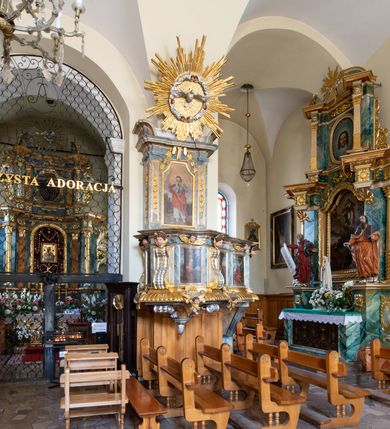 Zdjęcie nr 17: Świątynia otoczona jest murem kamiennym, tynkowanym, nakrytym daszkiem gontowym, jednospadowym. Wejścia znajdują się w murze od strony południowej. Pierwsze, prowadzące do głównych drzwi jest kamienną, późnobarokową bramką z herbem księdza Józefa Sawickiego, datą „1765” oraz figurą św. Jana Kantego. Drugie wejście prowadzi przez dzwonnicę. W murze, od strony wewnętrznej jest 14 wnęk po stacjach drogi krzyżowej. Teren wokół świątyni jest starym cmentarzem, bez zachowanych nagrobków.

Kościół jest budowlą założoną na planie prostokąta, orientowaną, jednonawową z ramionami w rodzaju krótkiego transeptu z wieżą od zachodu, zakrystią i kaplicą od północy oraz kruchtą od południa. Prezbiterium zamknięte jest wielobocznie, trójprzęsłowe, węższe i niższe od nawy. Nawa czteroprzęsłowa, nie jest ulokowana na jednej osi z prezbiterium. 

Artykulacja ścian jest przeprowadzona za pomocą pilastrów toskańskich podtrzymujących sklepienie kolebkowe z lunetami. Ramiona transeptu, kaplica Matki Boskiej Różańcowej oraz kruchta są sklepione krzyżowo. Otwory okienne: w prezbiterium cztery, w nawie cztery od południa, wszystkie zamknięte łukami półkolistymi. Zakrystia piętrowa z jednym oknem na parterze i dwoma na piętrze; od strony północnej kaplica Matki Boskiej Różańcowej z dwoma oknami i jedną, zamurowaną wnęką; obok mała zakrystia. Z prezbiterium do zakrystii prowadzi portal kamienny z inicjałami, herbem i portretem księdza Sawickiego. Na arkadzie tęczowej od strony nawy nowy napis: „FUNDATA A CASIMIRO MAGNO ANNO DOM[INI] 1346”. W pierwszym od strony fasady przęśle znajduje się drewniany chór muzyczny, wsparty na dwóch filarach.

Fasada zachodnia kościoła jest płaska z wieżą, nakryta kopułą z latarnią. Na narożnikach zewnętrznych wieży i naw oraz przy absydzie szkarpy, kryte daszkiem gontowym. Na frontowej ścianie wieży kamienny portal, nad nim sygle „I(osephus) S(awicki) / 1765” oraz herb Nowina. Od strony zachodniej wieża, ozdobiona półkolistymi blendami. Od strony południowej balkonik otoczony żelazną kratą z napisem „1343-1943”. Nad balkonikiem umieszczona jest tarcza zegara, taka sama widnieje od strony północnej, a od zachodniej i wschodniej odpowiadają im tarcze w murze. Przy wieży, od strony północnej wieżyczka w kształcie walca, ścięta od połowy, nakryta daszkiem stożkowym. Przy południowej ścianie wieży między dwiema szkarpami znajduje się kapliczka Ukrzyżowania z figurami Chrystusa Ukrzyżowanego i dwóch łotrów na krzyżach. Dach kościoła siodłowy, kryty gontem.

Na zewnętrznej, południowej ścianie napis: „AEDIFICATA H(A)EC EC[C]LESIA / A[NNO] D[OMINI] / 1606 / PETRVS CZARNY MVLARS”. Niżej wmurowana w półokrągłą wnękę z piaskowca figura Matki Boskiej Niepokalanie Poczętej. Po lewej stronie, u góry fragment fryzu dekoracyjnego, malowany w kolorze czerwonym o motywie zwisających esownic ze stylizowanymi kwiatami i liliami. 

Dzwonnica na planie kwadratu, murowana, otynkowana, dwupiętrowa z sienią przejściową sklepioną, ujętą na zaokrąglonych narożnikach zdwojonymi pilastrami; nakryta gontowym hełmem. Na osi dzwonnicy portal bramy flankowany dwoma pilastrami, o spłaszczonej archiwolcie. Nad belkowaniem monogram maryjny i data „1701”. W sieni na ścianie wschodniej barokowy krucyfiks oraz rzeźby Matki Boskiej i św. Jana Ewangelisty, na ścianie pozostałości polichromii z XVIII wieku.
