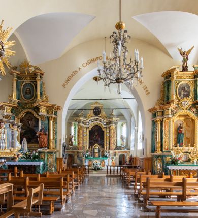 Zdjęcie nr 12: Świątynia otoczona jest murem kamiennym, tynkowanym, nakrytym daszkiem gontowym, jednospadowym. Wejścia znajdują się w murze od strony południowej. Pierwsze, prowadzące do głównych drzwi jest kamienną, późnobarokową bramką z herbem księdza Józefa Sawickiego, datą „1765” oraz figurą św. Jana Kantego. Drugie wejście prowadzi przez dzwonnicę. W murze, od strony wewnętrznej jest 14 wnęk po stacjach drogi krzyżowej. Teren wokół świątyni jest starym cmentarzem, bez zachowanych nagrobków.

Kościół jest budowlą założoną na planie prostokąta, orientowaną, jednonawową z ramionami w rodzaju krótkiego transeptu z wieżą od zachodu, zakrystią i kaplicą od północy oraz kruchtą od południa. Prezbiterium zamknięte jest wielobocznie, trójprzęsłowe, węższe i niższe od nawy. Nawa czteroprzęsłowa, nie jest ulokowana na jednej osi z prezbiterium. 

Artykulacja ścian jest przeprowadzona za pomocą pilastrów toskańskich podtrzymujących sklepienie kolebkowe z lunetami. Ramiona transeptu, kaplica Matki Boskiej Różańcowej oraz kruchta są sklepione krzyżowo. Otwory okienne: w prezbiterium cztery, w nawie cztery od południa, wszystkie zamknięte łukami półkolistymi. Zakrystia piętrowa z jednym oknem na parterze i dwoma na piętrze; od strony północnej kaplica Matki Boskiej Różańcowej z dwoma oknami i jedną, zamurowaną wnęką; obok mała zakrystia. Z prezbiterium do zakrystii prowadzi portal kamienny z inicjałami, herbem i portretem księdza Sawickiego. Na arkadzie tęczowej od strony nawy nowy napis: „FUNDATA A CASIMIRO MAGNO ANNO DOM[INI] 1346”. W pierwszym od strony fasady przęśle znajduje się drewniany chór muzyczny, wsparty na dwóch filarach.

Fasada zachodnia kościoła jest płaska z wieżą, nakryta kopułą z latarnią. Na narożnikach zewnętrznych wieży i naw oraz przy absydzie szkarpy, kryte daszkiem gontowym. Na frontowej ścianie wieży kamienny portal, nad nim sygle „I(osephus) S(awicki) / 1765” oraz herb Nowina. Od strony zachodniej wieża, ozdobiona półkolistymi blendami. Od strony południowej balkonik otoczony żelazną kratą z napisem „1343-1943”. Nad balkonikiem umieszczona jest tarcza zegara, taka sama widnieje od strony północnej, a od zachodniej i wschodniej odpowiadają im tarcze w murze. Przy wieży, od strony północnej wieżyczka w kształcie walca, ścięta od połowy, nakryta daszkiem stożkowym. Przy południowej ścianie wieży między dwiema szkarpami znajduje się kapliczka Ukrzyżowania z figurami Chrystusa Ukrzyżowanego i dwóch łotrów na krzyżach. Dach kościoła siodłowy, kryty gontem.

Na zewnętrznej, południowej ścianie napis: „AEDIFICATA H(A)EC EC[C]LESIA / A[NNO] D[OMINI] / 1606 / PETRVS CZARNY MVLARS”. Niżej wmurowana w półokrągłą wnękę z piaskowca figura Matki Boskiej Niepokalanie Poczętej. Po lewej stronie, u góry fragment fryzu dekoracyjnego, malowany w kolorze czerwonym o motywie zwisających esownic ze stylizowanymi kwiatami i liliami. 

Dzwonnica na planie kwadratu, murowana, otynkowana, dwupiętrowa z sienią przejściową sklepioną, ujętą na zaokrąglonych narożnikach zdwojonymi pilastrami; nakryta gontowym hełmem. Na osi dzwonnicy portal bramy flankowany dwoma pilastrami, o spłaszczonej archiwolcie. Nad belkowaniem monogram maryjny i data „1701”. W sieni na ścianie wschodniej barokowy krucyfiks oraz rzeźby Matki Boskiej i św. Jana Ewangelisty, na ścianie pozostałości polichromii z XVIII wieku.
