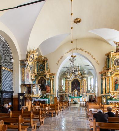 Zdjęcie nr 14: Świątynia otoczona jest murem kamiennym, tynkowanym, nakrytym daszkiem gontowym, jednospadowym. Wejścia znajdują się w murze od strony południowej. Pierwsze, prowadzące do głównych drzwi jest kamienną, późnobarokową bramką z herbem księdza Józefa Sawickiego, datą „1765” oraz figurą św. Jana Kantego. Drugie wejście prowadzi przez dzwonnicę. W murze, od strony wewnętrznej jest 14 wnęk po stacjach drogi krzyżowej. Teren wokół świątyni jest starym cmentarzem, bez zachowanych nagrobków.

Kościół jest budowlą założoną na planie prostokąta, orientowaną, jednonawową z ramionami w rodzaju krótkiego transeptu z wieżą od zachodu, zakrystią i kaplicą od północy oraz kruchtą od południa. Prezbiterium zamknięte jest wielobocznie, trójprzęsłowe, węższe i niższe od nawy. Nawa czteroprzęsłowa, nie jest ulokowana na jednej osi z prezbiterium. 

Artykulacja ścian jest przeprowadzona za pomocą pilastrów toskańskich podtrzymujących sklepienie kolebkowe z lunetami. Ramiona transeptu, kaplica Matki Boskiej Różańcowej oraz kruchta są sklepione krzyżowo. Otwory okienne: w prezbiterium cztery, w nawie cztery od południa, wszystkie zamknięte łukami półkolistymi. Zakrystia piętrowa z jednym oknem na parterze i dwoma na piętrze; od strony północnej kaplica Matki Boskiej Różańcowej z dwoma oknami i jedną, zamurowaną wnęką; obok mała zakrystia. Z prezbiterium do zakrystii prowadzi portal kamienny z inicjałami, herbem i portretem księdza Sawickiego. Na arkadzie tęczowej od strony nawy nowy napis: „FUNDATA A CASIMIRO MAGNO ANNO DOM[INI] 1346”. W pierwszym od strony fasady przęśle znajduje się drewniany chór muzyczny, wsparty na dwóch filarach.

Fasada zachodnia kościoła jest płaska z wieżą, nakryta kopułą z latarnią. Na narożnikach zewnętrznych wieży i naw oraz przy absydzie szkarpy, kryte daszkiem gontowym. Na frontowej ścianie wieży kamienny portal, nad nim sygle „I(osephus) S(awicki) / 1765” oraz herb Nowina. Od strony zachodniej wieża, ozdobiona półkolistymi blendami. Od strony południowej balkonik otoczony żelazną kratą z napisem „1343-1943”. Nad balkonikiem umieszczona jest tarcza zegara, taka sama widnieje od strony północnej, a od zachodniej i wschodniej odpowiadają im tarcze w murze. Przy wieży, od strony północnej wieżyczka w kształcie walca, ścięta od połowy, nakryta daszkiem stożkowym. Przy południowej ścianie wieży między dwiema szkarpami znajduje się kapliczka Ukrzyżowania z figurami Chrystusa Ukrzyżowanego i dwóch łotrów na krzyżach. Dach kościoła siodłowy, kryty gontem.

Na zewnętrznej, południowej ścianie napis: „AEDIFICATA H(A)EC EC[C]LESIA / A[NNO] D[OMINI] / 1606 / PETRVS CZARNY MVLARS”. Niżej wmurowana w półokrągłą wnękę z piaskowca figura Matki Boskiej Niepokalanie Poczętej. Po lewej stronie, u góry fragment fryzu dekoracyjnego, malowany w kolorze czerwonym o motywie zwisających esownic ze stylizowanymi kwiatami i liliami. 

Dzwonnica na planie kwadratu, murowana, otynkowana, dwupiętrowa z sienią przejściową sklepioną, ujętą na zaokrąglonych narożnikach zdwojonymi pilastrami; nakryta gontowym hełmem. Na osi dzwonnicy portal bramy flankowany dwoma pilastrami, o spłaszczonej archiwolcie. Nad belkowaniem monogram maryjny i data „1701”. W sieni na ścianie wschodniej barokowy krucyfiks oraz rzeźby Matki Boskiej i św. Jana Ewangelisty, na ścianie pozostałości polichromii z XVIII wieku.
