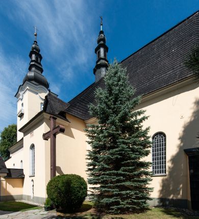 Zdjęcie nr 3: Świątynia otoczona jest murem kamiennym, tynkowanym, nakrytym daszkiem gontowym, jednospadowym. Wejścia znajdują się w murze od strony południowej. Pierwsze, prowadzące do głównych drzwi jest kamienną, późnobarokową bramką z herbem księdza Józefa Sawickiego, datą „1765” oraz figurą św. Jana Kantego. Drugie wejście prowadzi przez dzwonnicę. W murze, od strony wewnętrznej jest 14 wnęk po stacjach drogi krzyżowej. Teren wokół świątyni jest starym cmentarzem, bez zachowanych nagrobków.

Kościół jest budowlą założoną na planie prostokąta, orientowaną, jednonawową z ramionami w rodzaju krótkiego transeptu z wieżą od zachodu, zakrystią i kaplicą od północy oraz kruchtą od południa. Prezbiterium zamknięte jest wielobocznie, trójprzęsłowe, węższe i niższe od nawy. Nawa czteroprzęsłowa, nie jest ulokowana na jednej osi z prezbiterium. 

Artykulacja ścian jest przeprowadzona za pomocą pilastrów toskańskich podtrzymujących sklepienie kolebkowe z lunetami. Ramiona transeptu, kaplica Matki Boskiej Różańcowej oraz kruchta są sklepione krzyżowo. Otwory okienne: w prezbiterium cztery, w nawie cztery od południa, wszystkie zamknięte łukami półkolistymi. Zakrystia piętrowa z jednym oknem na parterze i dwoma na piętrze; od strony północnej kaplica Matki Boskiej Różańcowej z dwoma oknami i jedną, zamurowaną wnęką; obok mała zakrystia. Z prezbiterium do zakrystii prowadzi portal kamienny z inicjałami, herbem i portretem księdza Sawickiego. Na arkadzie tęczowej od strony nawy nowy napis: „FUNDATA A CASIMIRO MAGNO ANNO DOM[INI] 1346”. W pierwszym od strony fasady przęśle znajduje się drewniany chór muzyczny, wsparty na dwóch filarach.

Fasada zachodnia kościoła jest płaska z wieżą, nakryta kopułą z latarnią. Na narożnikach zewnętrznych wieży i naw oraz przy absydzie szkarpy, kryte daszkiem gontowym. Na frontowej ścianie wieży kamienny portal, nad nim sygle „I(osephus) S(awicki) / 1765” oraz herb Nowina. Od strony zachodniej wieża, ozdobiona półkolistymi blendami. Od strony południowej balkonik otoczony żelazną kratą z napisem „1343-1943”. Nad balkonikiem umieszczona jest tarcza zegara, taka sama widnieje od strony północnej, a od zachodniej i wschodniej odpowiadają im tarcze w murze. Przy wieży, od strony północnej wieżyczka w kształcie walca, ścięta od połowy, nakryta daszkiem stożkowym. Przy południowej ścianie wieży między dwiema szkarpami znajduje się kapliczka Ukrzyżowania z figurami Chrystusa Ukrzyżowanego i dwóch łotrów na krzyżach. Dach kościoła siodłowy, kryty gontem.

Na zewnętrznej, południowej ścianie napis: „AEDIFICATA H(A)EC EC[C]LESIA / A[NNO] D[OMINI] / 1606 / PETRVS CZARNY MVLARS”. Niżej wmurowana w półokrągłą wnękę z piaskowca figura Matki Boskiej Niepokalanie Poczętej. Po lewej stronie, u góry fragment fryzu dekoracyjnego, malowany w kolorze czerwonym o motywie zwisających esownic ze stylizowanymi kwiatami i liliami. 

Dzwonnica na planie kwadratu, murowana, otynkowana, dwupiętrowa z sienią przejściową sklepioną, ujętą na zaokrąglonych narożnikach zdwojonymi pilastrami; nakryta gontowym hełmem. Na osi dzwonnicy portal bramy flankowany dwoma pilastrami, o spłaszczonej archiwolcie. Nad belkowaniem monogram maryjny i data „1701”. W sieni na ścianie wschodniej barokowy krucyfiks oraz rzeźby Matki Boskiej i św. Jana Ewangelisty, na ścianie pozostałości polichromii z XVIII wieku.
