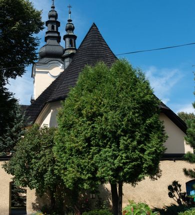 Zdjęcie nr 8: Świątynia otoczona jest murem kamiennym, tynkowanym, nakrytym daszkiem gontowym, jednospadowym. Wejścia znajdują się w murze od strony południowej. Pierwsze, prowadzące do głównych drzwi jest kamienną, późnobarokową bramką z herbem księdza Józefa Sawickiego, datą „1765” oraz figurą św. Jana Kantego. Drugie wejście prowadzi przez dzwonnicę. W murze, od strony wewnętrznej jest 14 wnęk po stacjach drogi krzyżowej. Teren wokół świątyni jest starym cmentarzem, bez zachowanych nagrobków.

Kościół jest budowlą założoną na planie prostokąta, orientowaną, jednonawową z ramionami w rodzaju krótkiego transeptu z wieżą od zachodu, zakrystią i kaplicą od północy oraz kruchtą od południa. Prezbiterium zamknięte jest wielobocznie, trójprzęsłowe, węższe i niższe od nawy. Nawa czteroprzęsłowa, nie jest ulokowana na jednej osi z prezbiterium. 

Artykulacja ścian jest przeprowadzona za pomocą pilastrów toskańskich podtrzymujących sklepienie kolebkowe z lunetami. Ramiona transeptu, kaplica Matki Boskiej Różańcowej oraz kruchta są sklepione krzyżowo. Otwory okienne: w prezbiterium cztery, w nawie cztery od południa, wszystkie zamknięte łukami półkolistymi. Zakrystia piętrowa z jednym oknem na parterze i dwoma na piętrze; od strony północnej kaplica Matki Boskiej Różańcowej z dwoma oknami i jedną, zamurowaną wnęką; obok mała zakrystia. Z prezbiterium do zakrystii prowadzi portal kamienny z inicjałami, herbem i portretem księdza Sawickiego. Na arkadzie tęczowej od strony nawy nowy napis: „FUNDATA A CASIMIRO MAGNO ANNO DOM[INI] 1346”. W pierwszym od strony fasady przęśle znajduje się drewniany chór muzyczny, wsparty na dwóch filarach.

Fasada zachodnia kościoła jest płaska z wieżą, nakryta kopułą z latarnią. Na narożnikach zewnętrznych wieży i naw oraz przy absydzie szkarpy, kryte daszkiem gontowym. Na frontowej ścianie wieży kamienny portal, nad nim sygle „I(osephus) S(awicki) / 1765” oraz herb Nowina. Od strony zachodniej wieża, ozdobiona półkolistymi blendami. Od strony południowej balkonik otoczony żelazną kratą z napisem „1343-1943”. Nad balkonikiem umieszczona jest tarcza zegara, taka sama widnieje od strony północnej, a od zachodniej i wschodniej odpowiadają im tarcze w murze. Przy wieży, od strony północnej wieżyczka w kształcie walca, ścięta od połowy, nakryta daszkiem stożkowym. Przy południowej ścianie wieży między dwiema szkarpami znajduje się kapliczka Ukrzyżowania z figurami Chrystusa Ukrzyżowanego i dwóch łotrów na krzyżach. Dach kościoła siodłowy, kryty gontem.

Na zewnętrznej, południowej ścianie napis: „AEDIFICATA H(A)EC EC[C]LESIA / A[NNO] D[OMINI] / 1606 / PETRVS CZARNY MVLARS”. Niżej wmurowana w półokrągłą wnękę z piaskowca figura Matki Boskiej Niepokalanie Poczętej. Po lewej stronie, u góry fragment fryzu dekoracyjnego, malowany w kolorze czerwonym o motywie zwisających esownic ze stylizowanymi kwiatami i liliami. 

Dzwonnica na planie kwadratu, murowana, otynkowana, dwupiętrowa z sienią przejściową sklepioną, ujętą na zaokrąglonych narożnikach zdwojonymi pilastrami; nakryta gontowym hełmem. Na osi dzwonnicy portal bramy flankowany dwoma pilastrami, o spłaszczonej archiwolcie. Nad belkowaniem monogram maryjny i data „1701”. W sieni na ścianie wschodniej barokowy krucyfiks oraz rzeźby Matki Boskiej i św. Jana Ewangelisty, na ścianie pozostałości polichromii z XVIII wieku.
