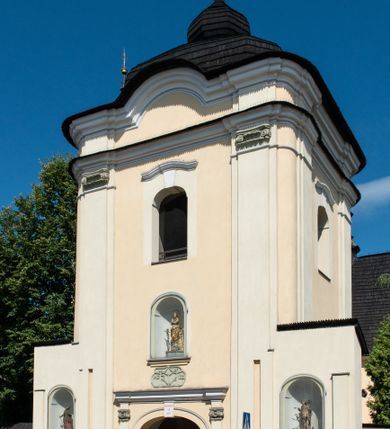 Zdjęcie nr 7: Świątynia otoczona jest murem kamiennym, tynkowanym, nakrytym daszkiem gontowym, jednospadowym. Wejścia znajdują się w murze od strony południowej. Pierwsze, prowadzące do głównych drzwi jest kamienną, późnobarokową bramką z herbem księdza Józefa Sawickiego, datą „1765” oraz figurą św. Jana Kantego. Drugie wejście prowadzi przez dzwonnicę. W murze, od strony wewnętrznej jest 14 wnęk po stacjach drogi krzyżowej. Teren wokół świątyni jest starym cmentarzem, bez zachowanych nagrobków.

Kościół jest budowlą założoną na planie prostokąta, orientowaną, jednonawową z ramionami w rodzaju krótkiego transeptu z wieżą od zachodu, zakrystią i kaplicą od północy oraz kruchtą od południa. Prezbiterium zamknięte jest wielobocznie, trójprzęsłowe, węższe i niższe od nawy. Nawa czteroprzęsłowa, nie jest ulokowana na jednej osi z prezbiterium. 

Artykulacja ścian jest przeprowadzona za pomocą pilastrów toskańskich podtrzymujących sklepienie kolebkowe z lunetami. Ramiona transeptu, kaplica Matki Boskiej Różańcowej oraz kruchta są sklepione krzyżowo. Otwory okienne: w prezbiterium cztery, w nawie cztery od południa, wszystkie zamknięte łukami półkolistymi. Zakrystia piętrowa z jednym oknem na parterze i dwoma na piętrze; od strony północnej kaplica Matki Boskiej Różańcowej z dwoma oknami i jedną, zamurowaną wnęką; obok mała zakrystia. Z prezbiterium do zakrystii prowadzi portal kamienny z inicjałami, herbem i portretem księdza Sawickiego. Na arkadzie tęczowej od strony nawy nowy napis: „FUNDATA A CASIMIRO MAGNO ANNO DOM[INI] 1346”. W pierwszym od strony fasady przęśle znajduje się drewniany chór muzyczny, wsparty na dwóch filarach.

Fasada zachodnia kościoła jest płaska z wieżą, nakryta kopułą z latarnią. Na narożnikach zewnętrznych wieży i naw oraz przy absydzie szkarpy, kryte daszkiem gontowym. Na frontowej ścianie wieży kamienny portal, nad nim sygle „I(osephus) S(awicki) / 1765” oraz herb Nowina. Od strony zachodniej wieża, ozdobiona półkolistymi blendami. Od strony południowej balkonik otoczony żelazną kratą z napisem „1343-1943”. Nad balkonikiem umieszczona jest tarcza zegara, taka sama widnieje od strony północnej, a od zachodniej i wschodniej odpowiadają im tarcze w murze. Przy wieży, od strony północnej wieżyczka w kształcie walca, ścięta od połowy, nakryta daszkiem stożkowym. Przy południowej ścianie wieży między dwiema szkarpami znajduje się kapliczka Ukrzyżowania z figurami Chrystusa Ukrzyżowanego i dwóch łotrów na krzyżach. Dach kościoła siodłowy, kryty gontem.

Na zewnętrznej, południowej ścianie napis: „AEDIFICATA H(A)EC EC[C]LESIA / A[NNO] D[OMINI] / 1606 / PETRVS CZARNY MVLARS”. Niżej wmurowana w półokrągłą wnękę z piaskowca figura Matki Boskiej Niepokalanie Poczętej. Po lewej stronie, u góry fragment fryzu dekoracyjnego, malowany w kolorze czerwonym o motywie zwisających esownic ze stylizowanymi kwiatami i liliami. 

Dzwonnica na planie kwadratu, murowana, otynkowana, dwupiętrowa z sienią przejściową sklepioną, ujętą na zaokrąglonych narożnikach zdwojonymi pilastrami; nakryta gontowym hełmem. Na osi dzwonnicy portal bramy flankowany dwoma pilastrami, o spłaszczonej archiwolcie. Nad belkowaniem monogram maryjny i data „1701”. W sieni na ścianie wschodniej barokowy krucyfiks oraz rzeźby Matki Boskiej i św. Jana Ewangelisty, na ścianie pozostałości polichromii z XVIII wieku.
