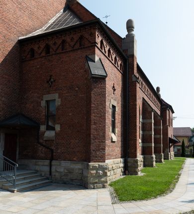 Zdjęcie nr 24: Kościół św. Jana Chrzciciela w Zembrzycach jest usytuowany na zachód od głównej drogi prowadzącej z Makowa Podhalańskiego do Wadowic i położony w centrum wsi, na placu otoczonym żeliwnym płotem na kamiennej podmurówce. Wejście na plac przykościelny prowadzi przez bramę od zachodu i wąską bramkę od północy. Kościół jest budowlą orientowaną, założoną na planie prostokąta, trójnawową, pseudohalową z prezbiterium zamkniętym trójbocznie, przy którym od północy znajduje się kaplica, a od południa piętrowa zakrystia z przedsionkiem i emporą otwartą na prezbiterium. Do nawy od strony północnej przylega wąska kruchta, nakryta dwuspadowym daszkiem. Od zachodu świątyni jest czworoboczna, masywna wieża, w górnej jednej trzeciej wysokości ośmioboczna, nakryta hełmem baniastym z latarnią. W fasadzie wysoka, półkolista wnęka z portalem i oknem termalnym. Przy wieży od północny składzik, a od południa kaplica. Ściany prezbiterium, nawy oraz elewacje aneksów w narożnikach artykułowane są szkarpami. Elewacje boczne zwieńczone ceglanym, zygzakowatym fryzem z krzyżykami. Szczyty na zakończeniach korpusu ozdobione kamiennymi szyszkami. W nawie okna trójdzielne z kamiennymi przezroczami, zamknięte łukami półkolistymi; w prezbiterium prostokątne zamknięte półkoliście. Świątynia nakryta jest dachem dwuspadowym. Na zakończeniu nawy znajduje się wieżyczka na sygnaturkę z baniastym hełmem. Wewnątrz nawy oddzielone są kolumnami z ośmiobocznymi głowicami, które wspierają sklepienia kolebkowe z lunetami. Empory i kaplice otwarte są arkadami półkolistymi. Prezbiterium podniesione jest o poziom dwóch stopni.
