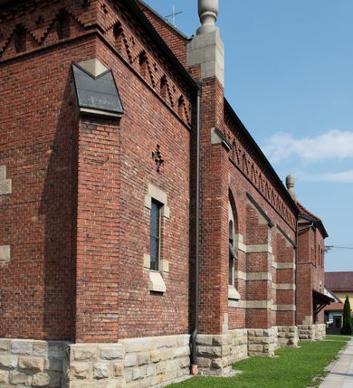 Zdjęcie nr 26: Kościół św. Jana Chrzciciela w Zembrzycach jest usytuowany na zachód od głównej drogi prowadzącej z Makowa Podhalańskiego do Wadowic i położony w centrum wsi, na placu otoczonym żeliwnym płotem na kamiennej podmurówce. Wejście na plac przykościelny prowadzi przez bramę od zachodu i wąską bramkę od północy. Kościół jest budowlą orientowaną, założoną na planie prostokąta, trójnawową, pseudohalową z prezbiterium zamkniętym trójbocznie, przy którym od północy znajduje się kaplica, a od południa piętrowa zakrystia z przedsionkiem i emporą otwartą na prezbiterium. Do nawy od strony północnej przylega wąska kruchta, nakryta dwuspadowym daszkiem. Od zachodu świątyni jest czworoboczna, masywna wieża, w górnej jednej trzeciej wysokości ośmioboczna, nakryta hełmem baniastym z latarnią. W fasadzie wysoka, półkolista wnęka z portalem i oknem termalnym. Przy wieży od północny składzik, a od południa kaplica. Ściany prezbiterium, nawy oraz elewacje aneksów w narożnikach artykułowane są szkarpami. Elewacje boczne zwieńczone ceglanym, zygzakowatym fryzem z krzyżykami. Szczyty na zakończeniach korpusu ozdobione kamiennymi szyszkami. W nawie okna trójdzielne z kamiennymi przezroczami, zamknięte łukami półkolistymi; w prezbiterium prostokątne zamknięte półkoliście. Świątynia nakryta jest dachem dwuspadowym. Na zakończeniu nawy znajduje się wieżyczka na sygnaturkę z baniastym hełmem. Wewnątrz nawy oddzielone są kolumnami z ośmiobocznymi głowicami, które wspierają sklepienia kolebkowe z lunetami. Empory i kaplice otwarte są arkadami półkolistymi. Prezbiterium podniesione jest o poziom dwóch stopni.
