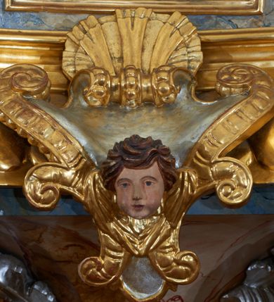 Zdjęcie nr 11: Ambona przyścienna, wisząca, z jednobiegowymi schodami dostępnymi od prawej strony. Kosz na planie kwadratu o ściętych narożach i wklęsło-wypukłym profilu. U spodu ośmioboczne podwieszenie zwężające się ku dołowi, ujęte parą spływów wolutowych utworzonych z liści akantu, u dołu zakończone szyszką. Cokół kosza zdobiony puklowaniem, dekorowany trzema kartuszami utworzonymi z karbowanej wstęgi, zwieńczonymi płaskimi, symetrycznymi palmetami, z główkami aniołków skierowanymi na wprost i umieszczonymi u dołu. Naroża kosza ozdobione spływami wolutowymi o karbowanych trzonach, ozdobionych kampanulami, podtrzymującymi belkowanie, stanowiące parapet z silnie wysuniętym gzymsem. Na fryzie parapetu uskrzydlone główki anielskie.

Ściany dekorowane są płycinami w formie stojących prostokątów, naprzemiennie wybrzuszonych lub wklęsłych z przedstawieniami czterech ewangelistów, ujętymi w złocone ramy. Od lewej: św. Mateusz, św. Marek, św. Łukasz i św. Jan. Święty Mateusz siedzi zwrócony trzy czwarte w lewo i pisze piórem w księdze. Obok stoi uskrzydlony człowiek, który obejmuje go za szyję, ubrany w niebieską suknię odsłaniającą klatkę piersiową i złoty płaszcz z końcem rozwianym ku górze. Święty ma rysy twarzy starszego człowieka z długą i siwą brodą oraz siwymi włosami. Ubrany jest w obszerny czerwony płaszcz i sandały. Scena ukazana jest we wnętrzu architektonicznym. Święty Marek również został ukazany w swojej pracowni. Siedzi, zwrócony trzy czwarte w lewo przy stoliku i pisząc w księdze, odwraca się w prawo, gdzie spoczywa lew – jego atrybut. Ma siwą brodę. Ubrany jest w niebieską suknię, zapinaną z przodu na guziki oraz czerwony płaszcz przewieszony przez prawe ramię. Stolik zasłany jest jasnozielonym obrusem, na którym leży otwarta księga i czerwony kałamarz. Na ścianie, ponad stolikiem wisi półka z książkami. Po lewej stronie obrazu widać otwartą przestrzeń z widokiem na góry, w pozostałej brązowe ściany pokoju, w którym znajduje się święty. Na kolejnym obrazie św. Łukasz siedzi zwrócony trzy czwarte w prawo, przy stoliku z pulpitem i o wolutowych nóżkach. W prawej dłoni trzyma pióro, którym pisze na karcie papieru. Ubrany jest z jasnobrązową suknię, niebieski płaszcz owijający nogi i niebieskie sandały; na głowie ma założoną czapkę. Po prawej stronie obrazu stoi jego atrybut – wół. Całość ukazana jest we wnętrzu architektonicznym z półkolistą arkadą po prawej stronie. Święty Jan został ukazany na tle krajobrazu, zwrócony jest trzy czwarte w prawo, pisze na karcie papieru. Ubrany jest w jasnozieloną suknię i czerwony płaszcz. Ma długie włosy, spływające na plecy, został ukazany bez zarostu. Po prawej stronie wysokie drzewo, w oddali pejzaż górski. Wszyscy ewangeliści mają okrągłe nimby z wpisaną w polu gwiazdą. 

Zaplecek w formie stojącego prostopadłościanu z niepełnym belkowaniem, wspartym na podwieszonych impostach, podtrzymującym odwrócony, przerywany przyczółek. Na ściance frontowej płycina w kształcie stojącego prostokąta o wklęsłych górnych narożach z obrazem Dobrego Pasterza w polu. Po bokach flankowana dwiema wąskimi płycinami, zdobionymi zwisem z kampanul. Na ściankach bocznych analogiczne płyciny z kampanulami. Chrystus ukazany frontalnie z barankiem na barkach, którego przytrzymuje rękami za nóżki. Zbawiciel ma owalną twarz z krótkim zarostem i jasnobrązowymi włosami, spływającymi na lewe ramię. Ubrany jest w biało-niebieską suknię oraz czerwony płaszcz. W oddali pejzaż z miastem po lewej, u góry szaro-złote niebo. Po lewej u dołu sygnatura „A D L”. Całość zwieńczona promienistą glorią z gołębicą Ducha Świętego w otoku utworzonym z chmur i główek anielskich. Struktura marmoryzowana w kolorze niebieskim, a płyciny w kolorze jasnoróżowym; ornamentyka, gloria i profile złocone i srebrzone.
