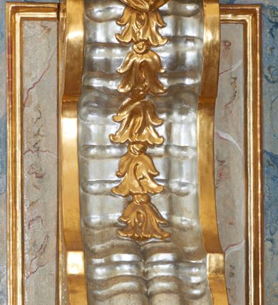 Zdjęcie nr 9: Ambona przyścienna, wisząca, z jednobiegowymi schodami dostępnymi od prawej strony. Kosz na planie kwadratu o ściętych narożach i wklęsło-wypukłym profilu. U spodu ośmioboczne podwieszenie zwężające się ku dołowi, ujęte parą spływów wolutowych utworzonych z liści akantu, u dołu zakończone szyszką. Cokół kosza zdobiony puklowaniem, dekorowany trzema kartuszami utworzonymi z karbowanej wstęgi, zwieńczonymi płaskimi, symetrycznymi palmetami, z główkami aniołków skierowanymi na wprost i umieszczonymi u dołu. Naroża kosza ozdobione spływami wolutowymi o karbowanych trzonach, ozdobionych kampanulami, podtrzymującymi belkowanie, stanowiące parapet z silnie wysuniętym gzymsem. Na fryzie parapetu uskrzydlone główki anielskie.

Ściany dekorowane są płycinami w formie stojących prostokątów, naprzemiennie wybrzuszonych lub wklęsłych z przedstawieniami czterech ewangelistów, ujętymi w złocone ramy. Od lewej: św. Mateusz, św. Marek, św. Łukasz i św. Jan. Święty Mateusz siedzi zwrócony trzy czwarte w lewo i pisze piórem w księdze. Obok stoi uskrzydlony człowiek, który obejmuje go za szyję, ubrany w niebieską suknię odsłaniającą klatkę piersiową i złoty płaszcz z końcem rozwianym ku górze. Święty ma rysy twarzy starszego człowieka z długą i siwą brodą oraz siwymi włosami. Ubrany jest w obszerny czerwony płaszcz i sandały. Scena ukazana jest we wnętrzu architektonicznym. Święty Marek również został ukazany w swojej pracowni. Siedzi, zwrócony trzy czwarte w lewo przy stoliku i pisząc w księdze, odwraca się w prawo, gdzie spoczywa lew – jego atrybut. Ma siwą brodę. Ubrany jest w niebieską suknię, zapinaną z przodu na guziki oraz czerwony płaszcz przewieszony przez prawe ramię. Stolik zasłany jest jasnozielonym obrusem, na którym leży otwarta księga i czerwony kałamarz. Na ścianie, ponad stolikiem wisi półka z książkami. Po lewej stronie obrazu widać otwartą przestrzeń z widokiem na góry, w pozostałej brązowe ściany pokoju, w którym znajduje się święty. Na kolejnym obrazie św. Łukasz siedzi zwrócony trzy czwarte w prawo, przy stoliku z pulpitem i o wolutowych nóżkach. W prawej dłoni trzyma pióro, którym pisze na karcie papieru. Ubrany jest z jasnobrązową suknię, niebieski płaszcz owijający nogi i niebieskie sandały; na głowie ma założoną czapkę. Po prawej stronie obrazu stoi jego atrybut – wół. Całość ukazana jest we wnętrzu architektonicznym z półkolistą arkadą po prawej stronie. Święty Jan został ukazany na tle krajobrazu, zwrócony jest trzy czwarte w prawo, pisze na karcie papieru. Ubrany jest w jasnozieloną suknię i czerwony płaszcz. Ma długie włosy, spływające na plecy, został ukazany bez zarostu. Po prawej stronie wysokie drzewo, w oddali pejzaż górski. Wszyscy ewangeliści mają okrągłe nimby z wpisaną w polu gwiazdą. 

Zaplecek w formie stojącego prostopadłościanu z niepełnym belkowaniem, wspartym na podwieszonych impostach, podtrzymującym odwrócony, przerywany przyczółek. Na ściance frontowej płycina w kształcie stojącego prostokąta o wklęsłych górnych narożach z obrazem Dobrego Pasterza w polu. Po bokach flankowana dwiema wąskimi płycinami, zdobionymi zwisem z kampanul. Na ściankach bocznych analogiczne płyciny z kampanulami. Chrystus ukazany frontalnie z barankiem na barkach, którego przytrzymuje rękami za nóżki. Zbawiciel ma owalną twarz z krótkim zarostem i jasnobrązowymi włosami, spływającymi na lewe ramię. Ubrany jest w biało-niebieską suknię oraz czerwony płaszcz. W oddali pejzaż z miastem po lewej, u góry szaro-złote niebo. Po lewej u dołu sygnatura „A D L”. Całość zwieńczona promienistą glorią z gołębicą Ducha Świętego w otoku utworzonym z chmur i główek anielskich. Struktura marmoryzowana w kolorze niebieskim, a płyciny w kolorze jasnoróżowym; ornamentyka, gloria i profile złocone i srebrzone.
