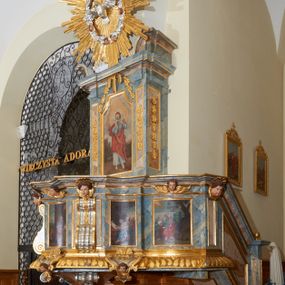 Zdjęcie nr 1: Ambona przyścienna, wisząca, z jednobiegowymi schodami dostępnymi od prawej strony. Kosz na planie kwadratu o ściętych narożach i wklęsło-wypukłym profilu. U spodu ośmioboczne podwieszenie zwężające się ku dołowi, ujęte parą spływów wolutowych utworzonych z liści akantu, u dołu zakończone szyszką. Cokół kosza zdobiony puklowaniem, dekorowany trzema kartuszami utworzonymi z karbowanej wstęgi, zwieńczonymi płaskimi, symetrycznymi palmetami, z główkami aniołków skierowanymi na wprost i umieszczonymi u dołu. Naroża kosza ozdobione spływami wolutowymi o karbowanych trzonach, ozdobionych kampanulami, podtrzymującymi belkowanie, stanowiące parapet z silnie wysuniętym gzymsem. Na fryzie parapetu uskrzydlone główki anielskie.

Ściany dekorowane są płycinami w formie stojących prostokątów, naprzemiennie wybrzuszonych lub wklęsłych z przedstawieniami czterech ewangelistów, ujętymi w złocone ramy. Od lewej: św. Mateusz, św. Marek, św. Łukasz i św. Jan. Święty Mateusz siedzi zwrócony trzy czwarte w lewo i pisze piórem w księdze. Obok stoi uskrzydlony człowiek, który obejmuje go za szyję, ubrany w niebieską suknię odsłaniającą klatkę piersiową i złoty płaszcz z końcem rozwianym ku górze. Święty ma rysy twarzy starszego człowieka z długą i siwą brodą oraz siwymi włosami. Ubrany jest w obszerny czerwony płaszcz i sandały. Scena ukazana jest we wnętrzu architektonicznym. Święty Marek również został ukazany w swojej pracowni. Siedzi, zwrócony trzy czwarte w lewo przy stoliku i pisząc w księdze, odwraca się w prawo, gdzie spoczywa lew – jego atrybut. Ma siwą brodę. Ubrany jest w niebieską suknię, zapinaną z przodu na guziki oraz czerwony płaszcz przewieszony przez prawe ramię. Stolik zasłany jest jasnozielonym obrusem, na którym leży otwarta księga i czerwony kałamarz. Na ścianie, ponad stolikiem wisi półka z książkami. Po lewej stronie obrazu widać otwartą przestrzeń z widokiem na góry, w pozostałej brązowe ściany pokoju, w którym znajduje się święty. Na kolejnym obrazie św. Łukasz siedzi zwrócony trzy czwarte w prawo, przy stoliku z pulpitem i o wolutowych nóżkach. W prawej dłoni trzyma pióro, którym pisze na karcie papieru. Ubrany jest z jasnobrązową suknię, niebieski płaszcz owijający nogi i niebieskie sandały; na głowie ma założoną czapkę. Po prawej stronie obrazu stoi jego atrybut – wół. Całość ukazana jest we wnętrzu architektonicznym z półkolistą arkadą po prawej stronie. Święty Jan został ukazany na tle krajobrazu, zwrócony jest trzy czwarte w prawo, pisze na karcie papieru. Ubrany jest w jasnozieloną suknię i czerwony płaszcz. Ma długie włosy, spływające na plecy, został ukazany bez zarostu. Po prawej stronie wysokie drzewo, w oddali pejzaż górski. Wszyscy ewangeliści mają okrągłe nimby z wpisaną w polu gwiazdą. 

Zaplecek w formie stojącego prostopadłościanu z niepełnym belkowaniem, wspartym na podwieszonych impostach, podtrzymującym odwrócony, przerywany przyczółek. Na ściance frontowej płycina w kształcie stojącego prostokąta o wklęsłych górnych narożach z obrazem Dobrego Pasterza w polu. Po bokach flankowana dwiema wąskimi płycinami, zdobionymi zwisem z kampanul. Na ściankach bocznych analogiczne płyciny z kampanulami. Chrystus ukazany frontalnie z barankiem na barkach, którego przytrzymuje rękami za nóżki. Zbawiciel ma owalną twarz z krótkim zarostem i jasnobrązowymi włosami, spływającymi na lewe ramię. Ubrany jest w biało-niebieską suknię oraz czerwony płaszcz. W oddali pejzaż z miastem po lewej, u góry szaro-złote niebo. Po lewej u dołu sygnatura „A D L”. Całość zwieńczona promienistą glorią z gołębicą Ducha Świętego w otoku utworzonym z chmur i główek anielskich. Struktura marmoryzowana w kolorze niebieskim, a płyciny w kolorze jasnoróżowym; ornamentyka, gloria i profile złocone i srebrzone.
