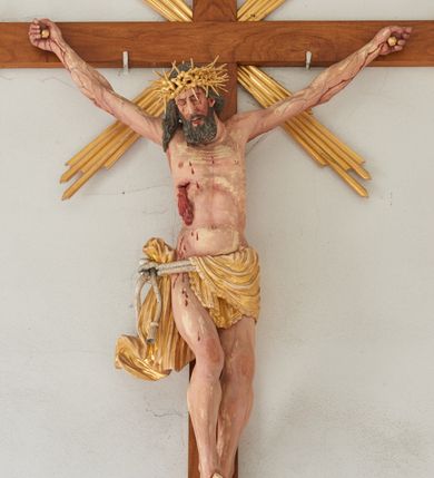 Zdjęcie nr 1: Chrystus przybity do krzyża na wyciągniętych do góry ramionach z prawą nogą ugiętą w kolanie i stopami przebitymi jednym gwoździem w układzie prawa na lewą. Klatka piersiowa smukła z wyraźnie zaznaczoną linią żeber i mostka, wciętą talią i dużą raną na prawym boku z wyrzeźbioną obficie broczącą krwią. Chrystus ma głowę opuszczoną w dół o podłużnej twarzy z zamkniętymi oczami, długim nosem i rozchylonymi ustami. Twarz okala bujna broda, rozdzielona na końcu w dwa szerokie pukle; włosy długie, ciemnobrązowe, opadają na prawe ramię. Perizonium jest przewiązane podwójnym sznurem, zakończonym chwostami. Tkanina na lewym boku Chrystusa i na środku jest przewieszona przez sznur i zdobiona na brzegach koronką, a po prawej stronie zawiązana u góry z końcem spływającym wzdłuż prawego uda, silnie drapowana, bez koronki. Na głowie Zbawiciela założona jest złocona, szeroka korona cierniowa z wbitymi oddzielnie długimi kolcami. Nad głową titulus w formie srebrzonej wstęgi z napisem „INRI”. Na skroniach, dłoniach i stopach ślady obficie spływającej krwi, na rękach Chrystusa wyraźnie zaznaczona siatka żył. Krzyż drewniany, gładki u góry i na bokach zakończony profilowanymi, złoconymi gzymsikami; od skrzyżowania ramion odchodzą cztery wiązki promieni o nierównej długości. Perizonium, korona cierniowa, wiązki promieni, gwoździe i gzymsiki złocone. 

