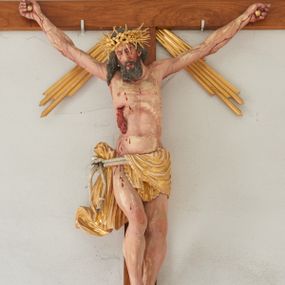 Zdjęcie nr 1: Chrystus przybity do krzyża na wyciągniętych do góry ramionach z prawą nogą ugiętą w kolanie i stopami przebitymi jednym gwoździem w układzie prawa na lewą. Klatka piersiowa smukła z wyraźnie zaznaczoną linią żeber i mostka, wciętą talią i dużą raną na prawym boku z wyrzeźbioną obficie broczącą krwią. Chrystus ma głowę opuszczoną w dół o podłużnej twarzy z zamkniętymi oczami, długim nosem i rozchylonymi ustami. Twarz okala bujna broda, rozdzielona na końcu w dwa szerokie pukle; włosy długie, ciemnobrązowe, opadają na prawe ramię. Perizonium jest przewiązane podwójnym sznurem, zakończonym chwostami. Tkanina na lewym boku Chrystusa i na środku jest przewieszona przez sznur i zdobiona na brzegach koronką, a po prawej stronie zawiązana u góry z końcem spływającym wzdłuż prawego uda, silnie drapowana, bez koronki. Na głowie Zbawiciela założona jest złocona, szeroka korona cierniowa z wbitymi oddzielnie długimi kolcami. Nad głową titulus w formie srebrzonej wstęgi z napisem „INRI”. Na skroniach, dłoniach i stopach ślady obficie spływającej krwi, na rękach Chrystusa wyraźnie zaznaczona siatka żył. Krzyż drewniany, gładki u góry i na bokach zakończony profilowanymi, złoconymi gzymsikami; od skrzyżowania ramion odchodzą cztery wiązki promieni o nierównej długości. Perizonium, korona cierniowa, wiązki promieni, gwoździe i gzymsiki złocone. 

