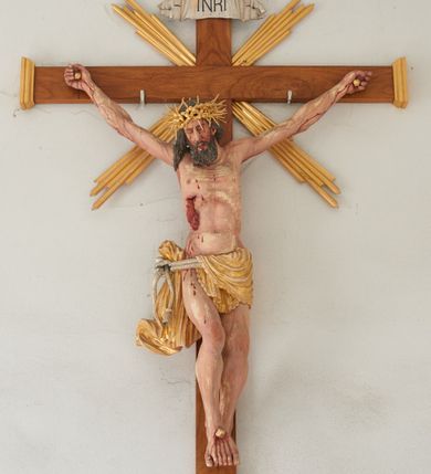 Zdjęcie nr 2: Chrystus przybity do krzyża na wyciągniętych do góry ramionach z prawą nogą ugiętą w kolanie i stopami przebitymi jednym gwoździem w układzie prawa na lewą. Klatka piersiowa smukła z wyraźnie zaznaczoną linią żeber i mostka, wciętą talią i dużą raną na prawym boku z wyrzeźbioną obficie broczącą krwią. Chrystus ma głowę opuszczoną w dół o podłużnej twarzy z zamkniętymi oczami, długim nosem i rozchylonymi ustami. Twarz okala bujna broda, rozdzielona na końcu w dwa szerokie pukle; włosy długie, ciemnobrązowe, opadają na prawe ramię. Perizonium jest przewiązane podwójnym sznurem, zakończonym chwostami. Tkanina na lewym boku Chrystusa i na środku jest przewieszona przez sznur i zdobiona na brzegach koronką, a po prawej stronie zawiązana u góry z końcem spływającym wzdłuż prawego uda, silnie drapowana, bez koronki. Na głowie Zbawiciela założona jest złocona, szeroka korona cierniowa z wbitymi oddzielnie długimi kolcami. Nad głową titulus w formie srebrzonej wstęgi z napisem „INRI”. Na skroniach, dłoniach i stopach ślady obficie spływającej krwi, na rękach Chrystusa wyraźnie zaznaczona siatka żył. Krzyż drewniany, gładki u góry i na bokach zakończony profilowanymi, złoconymi gzymsikami; od skrzyżowania ramion odchodzą cztery wiązki promieni o nierównej długości. Perizonium, korona cierniowa, wiązki promieni, gwoździe i gzymsiki złocone. 

