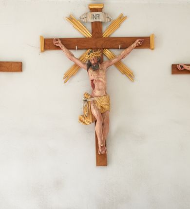 Zdjęcie nr 3: Chrystus przybity do krzyża na wyciągniętych do góry ramionach z prawą nogą ugiętą w kolanie i stopami przebitymi jednym gwoździem w układzie prawa na lewą. Klatka piersiowa smukła z wyraźnie zaznaczoną linią żeber i mostka, wciętą talią i dużą raną na prawym boku z wyrzeźbioną obficie broczącą krwią. Chrystus ma głowę opuszczoną w dół o podłużnej twarzy z zamkniętymi oczami, długim nosem i rozchylonymi ustami. Twarz okala bujna broda, rozdzielona na końcu w dwa szerokie pukle; włosy długie, ciemnobrązowe, opadają na prawe ramię. Perizonium jest przewiązane podwójnym sznurem, zakończonym chwostami. Tkanina na lewym boku Chrystusa i na środku jest przewieszona przez sznur i zdobiona na brzegach koronką, a po prawej stronie zawiązana u góry z końcem spływającym wzdłuż prawego uda, silnie drapowana, bez koronki. Na głowie Zbawiciela założona jest złocona, szeroka korona cierniowa z wbitymi oddzielnie długimi kolcami. Nad głową titulus w formie srebrzonej wstęgi z napisem „INRI”. Na skroniach, dłoniach i stopach ślady obficie spływającej krwi, na rękach Chrystusa wyraźnie zaznaczona siatka żył. Krzyż drewniany, gładki u góry i na bokach zakończony profilowanymi, złoconymi gzymsikami; od skrzyżowania ramion odchodzą cztery wiązki promieni o nierównej długości. Perizonium, korona cierniowa, wiązki promieni, gwoździe i gzymsiki złocone. 

