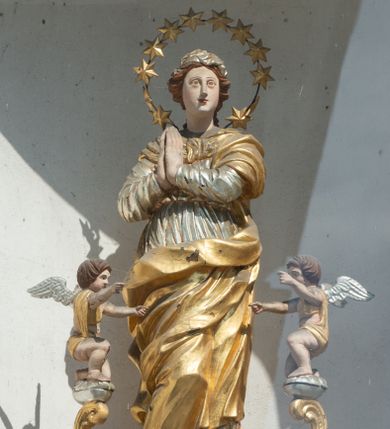 Zdjęcie nr 1: Figura Matki Boskiej Niepokalanie Poczętej umieszczona we wnęce na sześciobocznym cokole. Maria ukazana frontalnie, stojąca na zielonym półksiężycu, w kontrapoście, z prawą nogą ugiętą w kolanie, z rękami złożonymi w geście modlitwy. Twarz podłużna z małymi ustami, długim nosem i oczami skierowanymi na wprost, okolona bujnymi włosami, spiętymi z tyłu. Ubrana jest w ciasno przylegającą do ciała srebrzoną suknię, przewiązaną w talii, złocony płaszcz zasłaniający dekolt, lewe ramię i nogi. Na głowie ma założoną małą chustkę, a wokół niej duży nimb złożony z 12 gwiazd zawieszonych na metalowym pręcie. U dołu znajdują się dwie uskrzydlone główki aniołków w ekspresyjnych pozach. Całość flankują dwa pionowe spływy wolutowe utworzone z liści akantu, na których szczycie, na małych obłokach stoją dwa aniołki, zwrócone ku Matce Bożej. Aniołki przyklękają na jednym kolanie, ręce wyciągają przed siebie, mają szeroko rozłożone skrzydła. Twarze o rysach dziecięcych, okryte bujnymi, ciemnymi, średniej długości włosami, zasłaniającymi uszy. Ubrane są w złote przepaski, przerzucone jednym końcem przez ramię. Polichromia w odsłoniętych partiach ciała naturalistyczna, szaty i detale złocone i srebrzone, półksiężyc w kolorze zielonym, pokryty laserunkiem. Pod niszą z figurą znajduje się płaskorzeźbiony monogram maryjny z napisem „A(NNO) D(OMINI) 1701”.