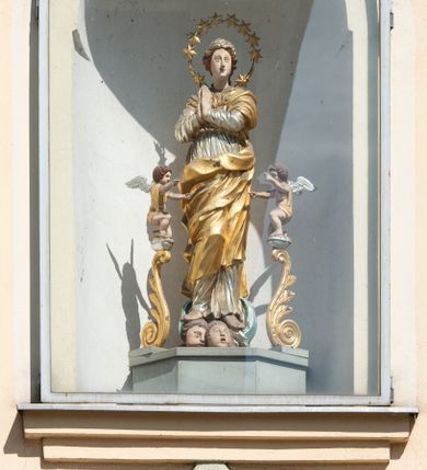 Zdjęcie nr 2: Figura Matki Boskiej Niepokalanie Poczętej umieszczona we wnęce na sześciobocznym cokole. Maria ukazana frontalnie, stojąca na zielonym półksiężycu, w kontrapoście, z prawą nogą ugiętą w kolanie, z rękami złożonymi w geście modlitwy. Twarz podłużna z małymi ustami, długim nosem i oczami skierowanymi na wprost, okolona bujnymi włosami, spiętymi z tyłu. Ubrana jest w ciasno przylegającą do ciała srebrzoną suknię, przewiązaną w talii, złocony płaszcz zasłaniający dekolt, lewe ramię i nogi. Na głowie ma założoną małą chustkę, a wokół niej duży nimb złożony z 12 gwiazd zawieszonych na metalowym pręcie. U dołu znajdują się dwie uskrzydlone główki aniołków w ekspresyjnych pozach. Całość flankują dwa pionowe spływy wolutowe utworzone z liści akantu, na których szczycie, na małych obłokach stoją dwa aniołki, zwrócone ku Matce Bożej. Aniołki przyklękają na jednym kolanie, ręce wyciągają przed siebie, mają szeroko rozłożone skrzydła. Twarze o rysach dziecięcych, okryte bujnymi, ciemnymi, średniej długości włosami, zasłaniającymi uszy. Ubrane są w złote przepaski, przerzucone jednym końcem przez ramię. Polichromia w odsłoniętych partiach ciała naturalistyczna, szaty i detale złocone i srebrzone, półksiężyc w kolorze zielonym, pokryty laserunkiem. Pod niszą z figurą znajduje się płaskorzeźbiony monogram maryjny z napisem „A(NNO) D(OMINI) 1701”.