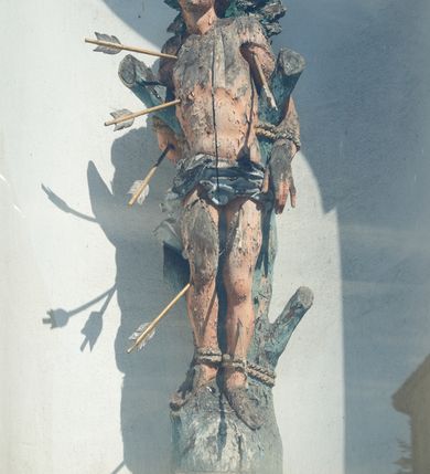 Zdjęcie nr 1: Rzeźba św. Sebastiana ustawiona na murowanym, wysokim cokole, zakończonym gzymsem. Święty przywiązany do pnia w postawie stojącej o delikatnie esowato wygiętej sylwetce, z rękami i nogami skrępowanymi sznurami, głowa przechylona w jego lewą stronę. Twarz o młodzieńczych rysach, pełna, usta drobne i otwarte, nos krótki i prosty, oczy podłużne. Włosy ciemnobrązowe, średniej długości, zasłaniające uszy, zaczesane do tyłu bujnymi lokami. Ciało owinięte w biodrach jasną przepaską. Święty ugodzony jest sześcioma strzałami. Polichromia w odsłoniętych partiach ciała naturalistyczna, strzały i detale złocone i srebrzone. 
