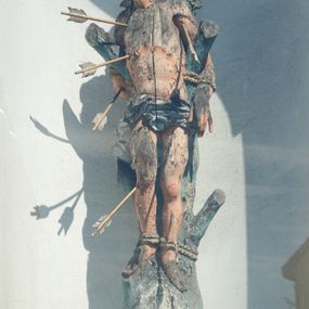 Zdjęcie nr 1: Rzeźba św. Sebastiana ustawiona na murowanym, wysokim cokole, zakończonym gzymsem. Święty przywiązany do pnia w postawie stojącej o delikatnie esowato wygiętej sylwetce, z rękami i nogami skrępowanymi sznurami, głowa przechylona w jego lewą stronę. Twarz o młodzieńczych rysach, pełna, usta drobne i otwarte, nos krótki i prosty, oczy podłużne. Włosy ciemnobrązowe, średniej długości, zasłaniające uszy, zaczesane do tyłu bujnymi lokami. Ciało owinięte w biodrach jasną przepaską. Święty ugodzony jest sześcioma strzałami. Polichromia w odsłoniętych partiach ciała naturalistyczna, strzały i detale złocone i srebrzone. 
