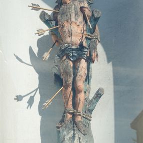 Zdjęcie nr 1: Rzeźba św. Sebastiana ustawiona na murowanym, wysokim cokole, zakończonym gzymsem. Święty przywiązany do pnia w postawie stojącej o delikatnie esowato wygiętej sylwetce, z rękami i nogami skrępowanymi sznurami, głowa przechylona w jego lewą stronę. Twarz o młodzieńczych rysach, pełna, usta drobne i otwarte, nos krótki i prosty, oczy podłużne. Włosy ciemnobrązowe, średniej długości, zasłaniające uszy, zaczesane do tyłu bujnymi lokami. Ciało owinięte w biodrach jasną przepaską. Święty ugodzony jest sześcioma strzałami. Polichromia w odsłoniętych partiach ciała naturalistyczna, strzały i detale złocone i srebrzone. 
