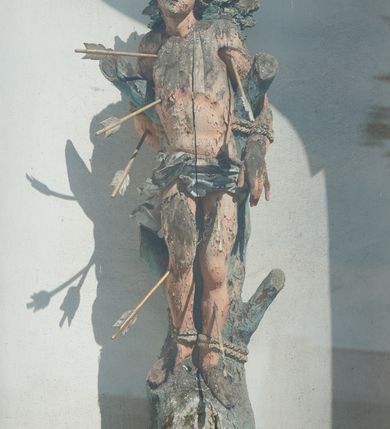 Zdjęcie nr 2: Rzeźba św. Sebastiana ustawiona na murowanym, wysokim cokole, zakończonym gzymsem. Święty przywiązany do pnia w postawie stojącej o delikatnie esowato wygiętej sylwetce, z rękami i nogami skrępowanymi sznurami, głowa przechylona w jego lewą stronę. Twarz o młodzieńczych rysach, pełna, usta drobne i otwarte, nos krótki i prosty, oczy podłużne. Włosy ciemnobrązowe, średniej długości, zasłaniające uszy, zaczesane do tyłu bujnymi lokami. Ciało owinięte w biodrach jasną przepaską. Święty ugodzony jest sześcioma strzałami. Polichromia w odsłoniętych partiach ciała naturalistyczna, strzały i detale złocone i srebrzone. 
