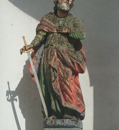 Zdjęcie nr 1: Rzeźba św. Pawła ustawiona jest na wysokim, drewnianym, prostopadłościennym cokole oraz niskiej kostce o ściętych narożach. Święty ukazany jest w postawie stojącej w silnym kontrapoście z lewą ręką złożoną na piersi i mieczem skierowanym ostrzem w dół w prawej dłoni. Święty głowę przechyla na lewe ramię, ma podłużną twarz z wyraźnie zaznaczonymi kośćmi policzkowymi, długim nosem i głęboko osadzonymi dużymi, oczami. Bujne i falowane włosy opadają na plecy i ramiona, a twarz okala gęsta broda. Apostoł ubrany jest w zieloną suknię z wąskimi rękawami oraz czerwony płaszcz zarzucony na prawe ramię i plecy z lewą połą przewieszoną z przodu przez pasek. Szaty drapowane twardo w ostre łamane fałdy. Święty ma małe, nieproporcjonalnie oddane dłonie w stosunku do całej sylwetki. Cokół polichromowany na niebiesko, miecz złocony i srebrzony.