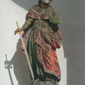 Zdjęcie nr 1: Rzeźba św. Pawła ustawiona jest na wysokim, drewnianym, prostopadłościennym cokole oraz niskiej kostce o ściętych narożach. Święty ukazany jest w postawie stojącej w silnym kontrapoście z lewą ręką złożoną na piersi i mieczem skierowanym ostrzem w dół w prawej dłoni. Święty głowę przechyla na lewe ramię, ma podłużną twarz z wyraźnie zaznaczonymi kośćmi policzkowymi, długim nosem i głęboko osadzonymi dużymi, oczami. Bujne i falowane włosy opadają na plecy i ramiona, a twarz okala gęsta broda. Apostoł ubrany jest w zieloną suknię z wąskimi rękawami oraz czerwony płaszcz zarzucony na prawe ramię i plecy z lewą połą przewieszoną z przodu przez pasek. Szaty drapowane twardo w ostre łamane fałdy. Święty ma małe, nieproporcjonalnie oddane dłonie w stosunku do całej sylwetki. Cokół polichromowany na niebiesko, miecz złocony i srebrzony.
