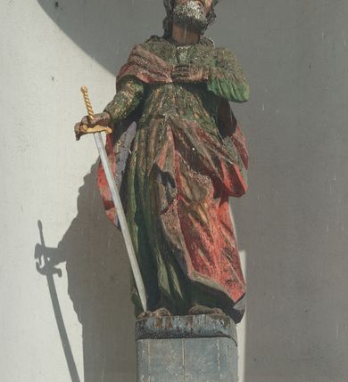 Zdjęcie nr 2: Rzeźba św. Pawła ustawiona jest na wysokim, drewnianym, prostopadłościennym cokole oraz niskiej kostce o ściętych narożach. Święty ukazany jest w postawie stojącej w silnym kontrapoście z lewą ręką złożoną na piersi i mieczem skierowanym ostrzem w dół w prawej dłoni. Święty głowę przechyla na lewe ramię, ma podłużną twarz z wyraźnie zaznaczonymi kośćmi policzkowymi, długim nosem i głęboko osadzonymi dużymi, oczami. Bujne i falowane włosy opadają na plecy i ramiona, a twarz okala gęsta broda. Apostoł ubrany jest w zieloną suknię z wąskimi rękawami oraz czerwony płaszcz zarzucony na prawe ramię i plecy z lewą połą przewieszoną z przodu przez pasek. Szaty drapowane twardo w ostre łamane fałdy. Święty ma małe, nieproporcjonalnie oddane dłonie w stosunku do całej sylwetki. Cokół polichromowany na niebiesko, miecz złocony i srebrzony.