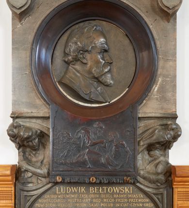 Zdjęcie nr 2: Epitafium w kształcie stojącego prostokąta, dwustopniowe, oddzielone wąskim gzymsem, zamknięte łukiem półkolistym, na którego zakończeniach dwie puste tarcze herbowe. W dolnej części epitafium płycina w kształcie leżącego prostokąta o fantazyjnym wykroju, w narożach ujęta dwiema czołgankami, z inskrypcją w polu: „LUDWIK BEŁTOWSKI / UR[ODZONY] 1819 WE LWOWIE ZASŁ[UŻONY] OBYW[ATEL] DŁUG[O]L[ETNI] RADNY MIASTA / NOWEGO TARGU MISTRZ ART[YSTA] BUD[OWLI] MECH[ANICZNYCH] PRZEM[YSŁOWYCH] PRZEWODN[ICZĄCY] / POW[IATOWEGO] STOW[ARZYSZENIA] RĘKODZ[IĘLNIKÓW] PRZEM[YSŁOWYCH] SĄDU POLUB[OWNEGO] INICIAT[OR] CZYNN[Y] ORĘD[OWNIK] / POWIĘKSZ[ANIA] MAJ[ĄTKU] DOBROBYTU GMINY UPIĘKSZ[ANIA] MIASTA OFIARN[Y] / PATRIOT[A] MĄŻ RZADK[ICH] CNÓT SERC[A] WIELK[IEGO] CHAR[AKTERU] ZM[ARŁ] 13. STYCZ[NIA] 1902 / PROSI O WESTCHNIENIE DO BOGA AMEN”. W prawym dolnym rogu epitafium sygnatura „Juliusz B[ełtowski] 1904.” Powyżej kwadratowy odlew z przedstawieniem Miłosiernego Samarytanina, flankowany dwiema popiersiami: rzeźbą Matki Boskiej z Dzieciątkiem po prawej i św. Józefa Robotnika po lewej. W dolnej części odlewu litery „D[EO] O[PTIMO] M[AXIMO]”. Na odlewie ukazany leżący na ziemi mężczyzna o wychudzonej posturze, któremu Samarytanin podaje kubek z wodą do picia. Po lewej stronie obrazu głowa wielbłąda. Matka Boska zwrócona trzy czwarte w lewo, z głową pochyloną w dół, przytulająca do piersi śpiące Dzieciątko. W dłoniach trzyma księgę. Twarz o wyraźnych rysach z długim nosem i zaciśniętymi ustami. Ubrana jest w suknię z długimi rękawami i płaszcz założony na głowę. Święty Józef zwrócony jest delikatnie w prawo z głową skierowaną w lewo. Ma krótkie i bujne włosy, wzrok skierowany w prawo oraz zaciśnięte usta. Ubrany jest w strój robotnika – koszulę z podwiniętymi do łokci rękawami. W dłoniach trzyma strug i piłę do drewna. W górnej części epitafium znajduje się okrągłe popiersie Ludwika Bełtowskiego w szerokiej okrągłej ramie. Zmarły ukazany z prawego profilu o podłużnej twarzy ze spiczastą brodą i wąsami, długim nosem i zmarszczonym czołem. Włosy krótkie. Ubrany jest w koszulę i marynarkę. Po lewej stronie popiersia sygnatura „B[ełtowski] [1]902”.



