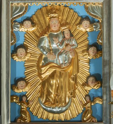 Zdjęcie nr 1: Obraz w kształcie stojącego prostokąta w drewnianej, srebrzonej ramie. W centrum kompozycji ukazana jest Matka Boska stojąca na półksiężycu z Dzieciątkiem Jezus na lewej ręce na tle złoconej, promienistej aureoli. Maria została ukazana w delikatnym kontrapoście z prawą nogą ugiętą w kolanie, w prawej dłoni trzyma berło. Ma szeroką, pełną twarz z dużym nosem oraz małymi ustami i oczami. Ubrana jest w srebrzoną suknię, przewiązaną w talii paskiem oraz złocony welon i złocony płaszcz osłaniający jej nogi i plecy. Dzieciątko siedzi zwrócone trzy czwarte w prawo, w lewej dłoni trzyma jabłko królewskie, a prawą czyni gest błogosławieństwa. Ma szeroką twarz o analogicznych rysach do twarzy Matki. Głowę Dzieciątka okalają długie, ciemne i bujne włosy. Maria i Dzieciątko mają na głowie założone pełne korony. W dolnej części kompozycji klęczą dwa aniołki, zwrócone do środka z twarzami w kierunku widza, ze złożonymi w geście modlitwy dłońmi. Po bokach obrazu umieszczonych jest też sześć uskrzydlonych główek anielskich skierowanych na wprost o pełnych twarzach i ciemnych, średniej długości włosach. W górnych narożach obrazu podwieszona jest srebrzona kotara ze złoconą bordiurą. Tło jednolite, błękitne. 