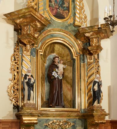 Zdjęcie nr 2: Ołtarz przyścienny, architektoniczny, jednoosiowy, jednokondygnacyjny ze zwieńczeniem. Mensa prostopadłościenna, w antepedium prostokątna, złocona rama. Prostokątna predella flankowana przed dwa wysunięte cokoły, na których ustawione są pilastry, wspierające przerwane belkowanie, zamknięte wydatnym, profilowanym gzymsem. Przed podporami ustawione są dwie figury świętych: po lewej stronie św. Teresy, a po prawej św. Kunegundy. W polu środkowym wnęka w kształcie stojącego prostokąta, zamkniętego łukiem nadwieszonym, w której umieszczona jest figura św. Antoniego Padewskiego na tle mniejszej płyciny o analogicznym kształcie. Imposty w partii fryzu ustawione są pod kątem, zdobione podwieszonymi od spodu szyszkami. Zwieńczenie w kształcie stojącego prostokąta, ujęte parą pilastrów, wspierających wyłamane belkowanie, zamknięte nad polem środkowym łukiem nadwieszonym. W polu obraz „Wniebowzięcie Najświętszej Marii Panny” w kształcie stojącego prostokąta od dołu i od góry zamkniętego łukiem nadwieszonym. Na szczycie zwieńczenia promienista gloria z monogramem maryjnym na tle obłoków. Podpory ustawione są na niskich bazach o trzonach dekorowanych płaską, skośnie oplatającą je szeroką wstęgą i o kapitelach korynckich. Boki nastawy i zwieńczenia zdobią uszaki formowane z ażurowej dekoracji rocaille’owej. Całość struktury ołtarza pokryta marmoryzacją zielono-niebieską; na cokole, fryzie, we wnęce i w zwieńczeniu cześciowo w kolorze jasnozielonym z jasnobrązowo-białym żyłkowaniem. Detale oraz profilowania pozłacane i posrebrzane. 
