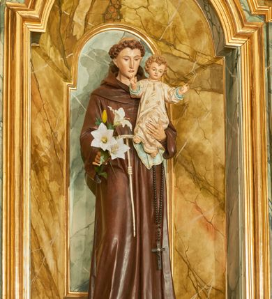 Zdjęcie nr 3: Rzeźba wolnostojąca z przedstawieniem św. Antoniego Padewskiego z Dzieciątkiem Jezus, ustawiona na cokole. Święty został ukazany w pozycji stojącej, w delikatnym kontrapoście z prawą nogą ugiętą w kolanie. Na lewej ręce trzyma Dzieciątko, a w prawej opuszczonej w dół gałązkę białej lilii (kwiaty sztuczne). Ma wąską twarz z długim nosem, drobne usta i małe oczy; na głowie tonsurę z silnie skręconych, krótkich włosów. Ubrany jest w brązowy habit franciszkański, przewiązany cingulum, a na lewym boku ma zawieszony długi, czarny różaniec; na stopach sandały. Cokół niski, prostopadłościenny o ściętych narożach, malowany w kolorze oliwkowozielonym. 