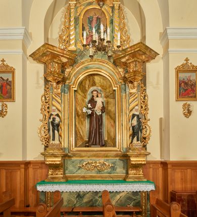 Zdjęcie nr 5: Rzeźba wolnostojąca z przedstawieniem św. Antoniego Padewskiego z Dzieciątkiem Jezus, ustawiona na cokole. Święty został ukazany w pozycji stojącej, w delikatnym kontrapoście z prawą nogą ugiętą w kolanie. Na lewej ręce trzyma Dzieciątko, a w prawej opuszczonej w dół gałązkę białej lilii (kwiaty sztuczne). Ma wąską twarz z długim nosem, drobne usta i małe oczy; na głowie tonsurę z silnie skręconych, krótkich włosów. Ubrany jest w brązowy habit franciszkański, przewiązany cingulum, a na lewym boku ma zawieszony długi, czarny różaniec; na stopach sandały. Cokół niski, prostopadłościenny o ściętych narożach, malowany w kolorze oliwkowozielonym. 
