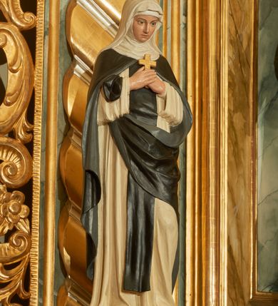 Zdjęcie nr 3: Rzeźba wolnostojąca z przedstawieniem św. Teresy Wielkiej, ustawiona na cokole. Święta została ukazana w pozycji stojącej, w delikatnym kontrapoście z lewą nogą ugiętą w kolanie. Zakonnica w skrzyżowanych na piersi dłoniach trzyma krzyżyk, a głowę pochyla delikatnie w dół. Ma owalną twarz z długim, szerokim nosem, drobne usta i duże oczy. Ubrana jest w biały habit, czarny szkaplerz, biały welon i podwikę oraz czarny płaszcz z lewą połą zarzuconą na prawe przedramię. Szaty drapowane są przeważnie w głębokie pionowe fałdy.  Cokół prostopadłościenny o ściętych narożach, marmoryzowany, na ściance przedniej wyryty napis: „S(ANCTA) TERESA.”