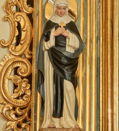 Zdjęcie nr 1: Rzeźba wolnostojąca z przedstawieniem św. Teresy Wielkiej, ustawiona na cokole. Święta została ukazana w pozycji stojącej, w delikatnym kontrapoście z lewą nogą ugiętą w kolanie. Zakonnica w skrzyżowanych na piersi dłoniach trzyma krzyżyk, a głowę pochyla delikatnie w dół. Ma owalną twarz z długim, szerokim nosem, drobne usta i duże oczy. Ubrana jest w biały habit, czarny szkaplerz, biały welon i podwikę oraz czarny płaszcz z lewą połą zarzuconą na prawe przedramię. Szaty drapowane są przeważnie w głębokie pionowe fałdy.  Cokół prostopadłościenny o ściętych narożach, marmoryzowany, na ściance przedniej wyryty napis: „S(ANCTA) TERESA.”