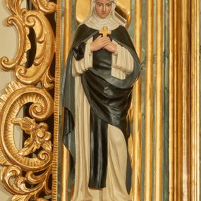 Zdjęcie nr 1: Rzeźba wolnostojąca z przedstawieniem św. Teresy Wielkiej, ustawiona na cokole. Święta została ukazana w pozycji stojącej, w delikatnym kontrapoście z lewą nogą ugiętą w kolanie. Zakonnica w skrzyżowanych na piersi dłoniach trzyma krzyżyk, a głowę pochyla delikatnie w dół. Ma owalną twarz z długim, szerokim nosem, drobne usta i duże oczy. Ubrana jest w biały habit, czarny szkaplerz, biały welon i podwikę oraz czarny płaszcz z lewą połą zarzuconą na prawe przedramię. Szaty drapowane są przeważnie w głębokie pionowe fałdy.  Cokół prostopadłościenny o ściętych narożach, marmoryzowany, na ściance przedniej wyryty napis: „S(ANCTA) TERESA.”