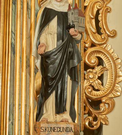 Zdjęcie nr 3: Rzeźba wolnostojąca z przedstawieniem św. Kunegundy, ustawiona na cokole. Święta została ukazana w pozycji stojącej, w delikatnym kontrapoście z prawą nogą ugiętą w kolanie. Zakonnica w lewej ręce trzyma model kościoła, a prawą opuszczoną w dół wyciąga przed siebie. Twarz owalna z długim i szerokim nosem, drobnymi ustami i dużymi oczami. Ubrana jest w biały habit, czarny szkaplerz, biały welon i podwikę oraz czarny płaszcz z prawą połą zarzuconą na lewe przedramię. Szaty drapowane są przeważnie w głębokie pionowe fałdy. Cokół prostopadłościenny o ściętych narożach, marmoryzowany, na ściance przedniej wyryty napis: „S(ANCTA) KUNEGUNDA”.