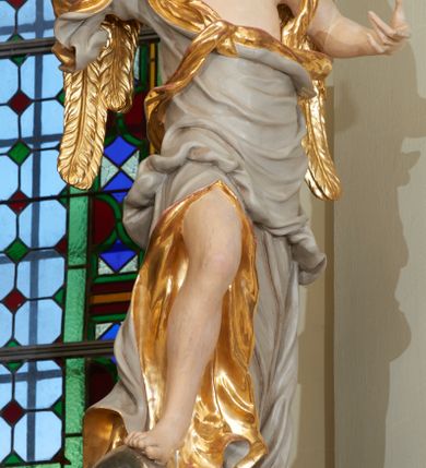 Zdjęcie nr 2: Rzeźba ścięta z tyłu, drążona. Figura ustawiona na kłębiącym się obłoku, zwrócona frontalnie, prawa noga uniesiona i oparta na obłoku, obie ręce wzniesione: prawa na piersi, lewa wysunięta w bok; głowa silnie przechylona na prawy bark. Twarz pociągła o wystającym podbródku, okolona rozwianymi, silnie skręconymi w pojedyncze pukle włosami. Anioł ubrany jest w szatę przerzuconą diagonalnie przez tors, odsłaniającą lewe ramię i rozchyloną w dolnej partii odkrywającą prawą nogę. Tkanina ściśle przylegająca, miejscami owinięta wokół ciała, silnie drapowana w falujące fałdy, w partii torsu z wywiniętym brzegiem, u dołu silnie rozwianym i zawijającym się fragmentarycznie. Skrzydła niewielkie, złożone. Polichromia naturalistyczna, szata w kolorze szarym ze złoconą podszewką, skrzydła złocone.
