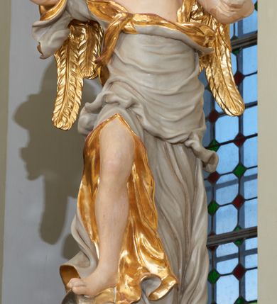 Zdjęcie nr 4: Rzeźba ścięta z tyłu, drążona. Figura ustawiona na kłębiącym się obłoku, zwrócona frontalnie, prawa noga uniesiona i oparta na obłoku, obie ręce wzniesione: prawa na piersi, lewa wysunięta w bok; głowa silnie przechylona na prawy bark. Twarz pociągła o wystającym podbródku, okolona rozwianymi, silnie skręconymi w pojedyncze pukle włosami. Anioł ubrany jest w szatę przerzuconą diagonalnie przez tors, odsłaniającą lewe ramię i rozchyloną w dolnej partii odkrywającą prawą nogę. Tkanina ściśle przylegająca, miejscami owinięta wokół ciała, silnie drapowana w falujące fałdy, w partii torsu z wywiniętym brzegiem, u dołu silnie rozwianym i zawijającym się fragmentarycznie. Skrzydła niewielkie, złożone. Polichromia naturalistyczna, szata w kolorze szarym ze złoconą podszewką, skrzydła złocone.
