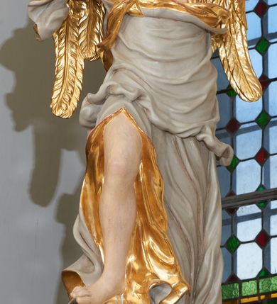 Zdjęcie nr 5: Rzeźba ścięta z tyłu, drążona. Figura ustawiona na kłębiącym się obłoku, zwrócona frontalnie, prawa noga uniesiona i oparta na obłoku, obie ręce wzniesione: prawa na piersi, lewa wysunięta w bok; głowa silnie przechylona na prawy bark. Twarz pociągła o wystającym podbródku, okolona rozwianymi, silnie skręconymi w pojedyncze pukle włosami. Anioł ubrany jest w szatę przerzuconą diagonalnie przez tors, odsłaniającą lewe ramię i rozchyloną w dolnej partii odkrywającą prawą nogę. Tkanina ściśle przylegająca, miejscami owinięta wokół ciała, silnie drapowana w falujące fałdy, w partii torsu z wywiniętym brzegiem, u dołu silnie rozwianym i zawijającym się fragmentarycznie. Skrzydła niewielkie, złożone. Polichromia naturalistyczna, szata w kolorze szarym ze złoconą podszewką, skrzydła złocone.
