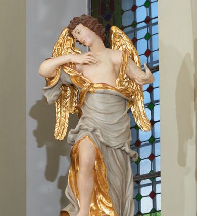 Zdjęcie nr 1: Rzeźba ścięta z tyłu, drążona. Figura ustawiona na kłębiącym się obłoku, zwrócona frontalnie, prawa noga uniesiona i oparta na obłoku, obie ręce wzniesione: prawa na piersi, lewa wysunięta w bok; głowa silnie przechylona na prawy bark. Twarz pociągła o wystającym podbródku, okolona rozwianymi, silnie skręconymi w pojedyncze pukle włosami. Anioł ubrany jest w szatę przerzuconą diagonalnie przez tors, odsłaniającą lewe ramię i rozchyloną w dolnej partii odkrywającą prawą nogę. Tkanina ściśle przylegająca, miejscami owinięta wokół ciała, silnie drapowana w falujące fałdy, w partii torsu z wywiniętym brzegiem, u dołu silnie rozwianym i zawijającym się fragmentarycznie. Skrzydła niewielkie, złożone. Polichromia naturalistyczna, szata w kolorze szarym ze złoconą podszewką, skrzydła złocone.
