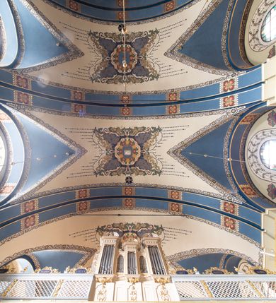 Zdjęcie nr 2: Dekoracja sklepienia nawy składa się z prostych symboli: kielicha z hostią, otwartej księgi z literami „A” i „Ω”, imienia MARIA i kotwicy. Zostały one wkomponowane w owalne medaliony w barwnej oprawie utworzonej z wieńców z żółtych i czerwonych kwiatów, złotych i zielonych wici akantów, ułożonych krzyżowo spiralnych, niebieskich ślimacznic i otaczających je stylizowanych zielonych liści akantu, między którymi przebijają się złote promienie. Owe fantazyjne kompozycje w nawie ukazane są na jasnym, kremowym tle wysklepek i niebieskich lunet i gurtów. Elementy konstrukcyjne szwów sklepiennych są podkreślone pasami z wici roślinnej. Sklepienie prezbiterium odróżnia się jaśniejszą kolorystyką. Elementy konstrukcyjne łuków podkreślają bujne czerwono-zielone kwiaty, wśród których rozpoznano piwonie. Taka sama bordiura zdobi ścianę za ołtarzem. Pośrodku umieszczono Baranka na zamkniętej księdze z chorągwią zmartwychwstania, na tle krzyża greckiego w glorii promienistej. Szwy sklepienne zaakcentowano kwiatami w układzie kandelabrowym, a wysklepki zielono-złotymi symetrycznie układającymi się wiciami roślinnymi. Na parapecie chóru muzycznego malarz przedstawił dwie sceny: „Stygmatyzacja św. Franciszka” i „Anioł grający na skrzypcach płaczącemu św. Franciszkowi”. 
